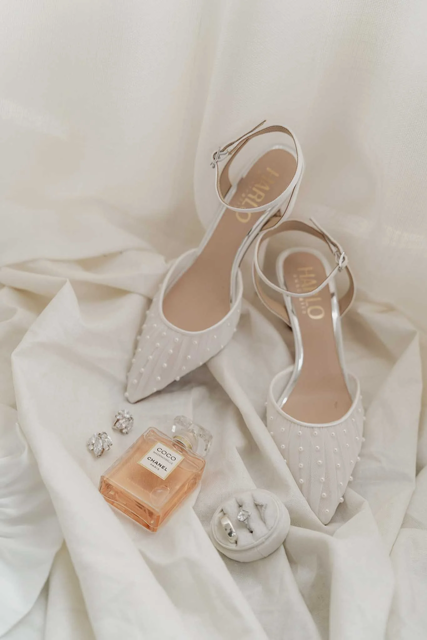 A pair of white high-heeled shoes with pearl embellishments, a bottle of Chanel Coco Mademoiselle perfume, diamond earrings, and a jewelry ring set are displayed on a white fabric background.