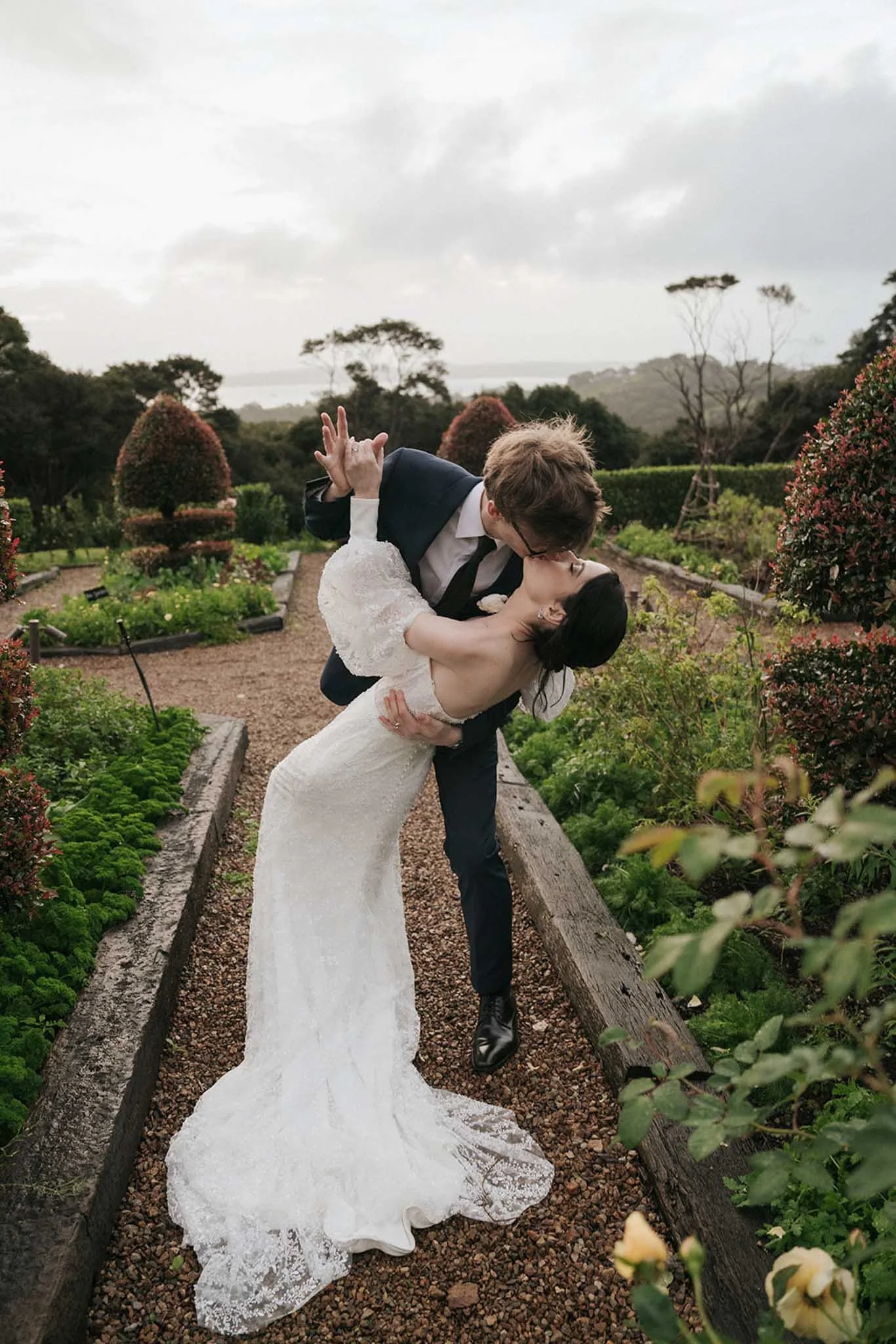 A groom dips and kisses a bride in a garden pathway surrounded by bushes and trees, with a cloudy sky overhead.