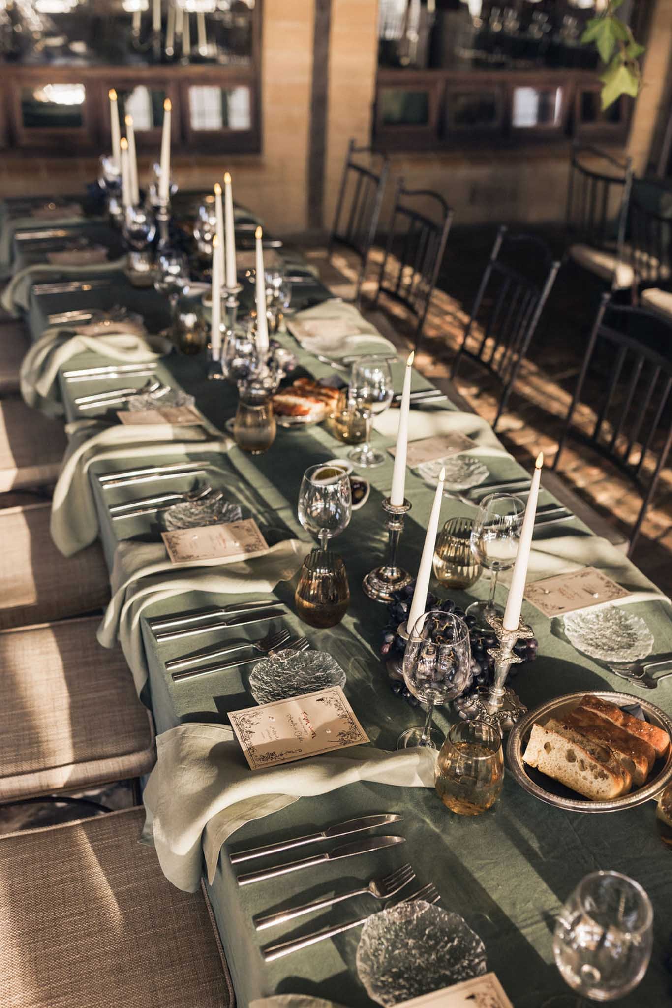 A long dining table set for a formal dinner, with white candles, wine glasses, plates, napkins, and a menu on a green tablecloth in a rustic room with brick walls and wooden window frames.