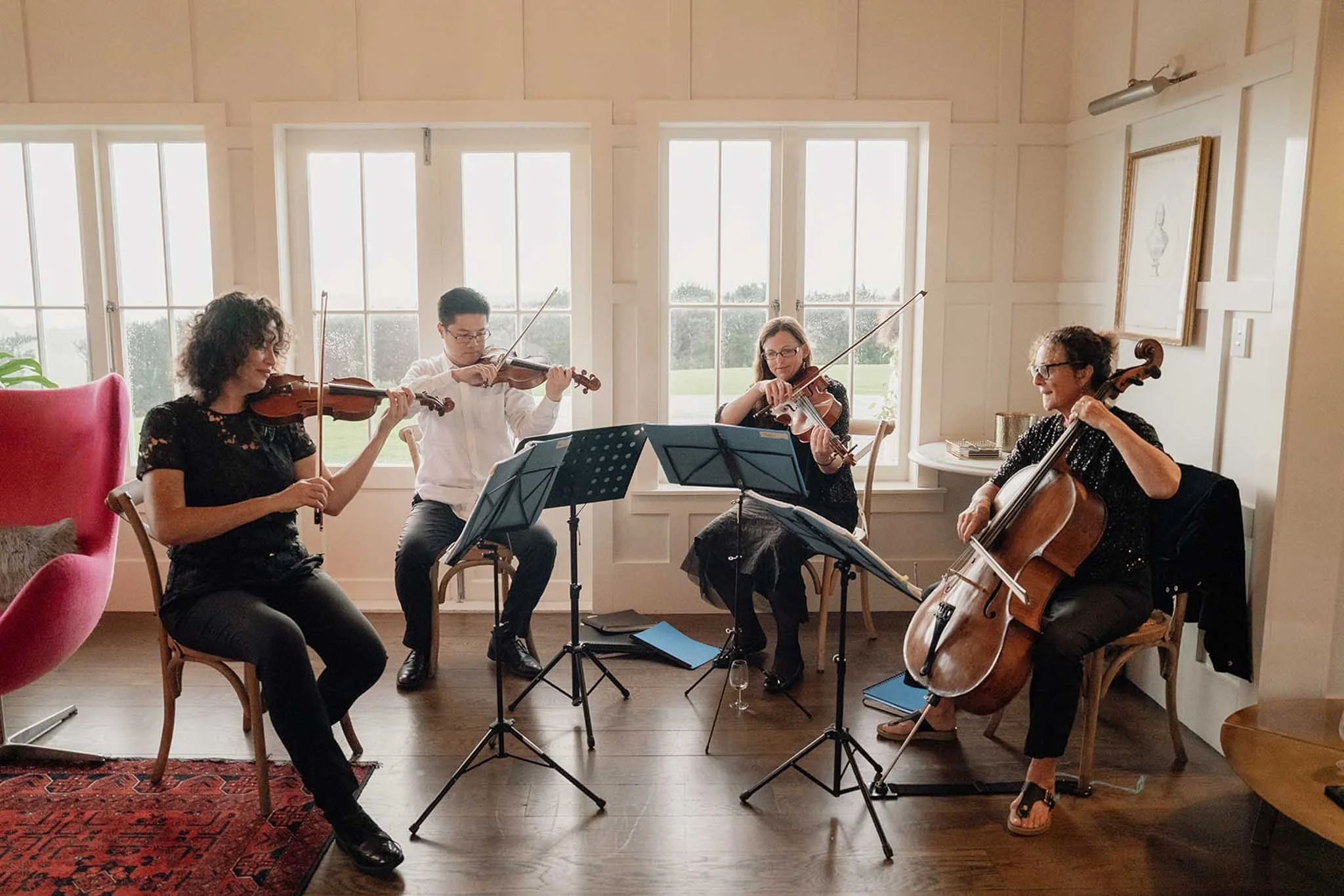 A group of five musicians playing string instruments in a bright room with large windows.