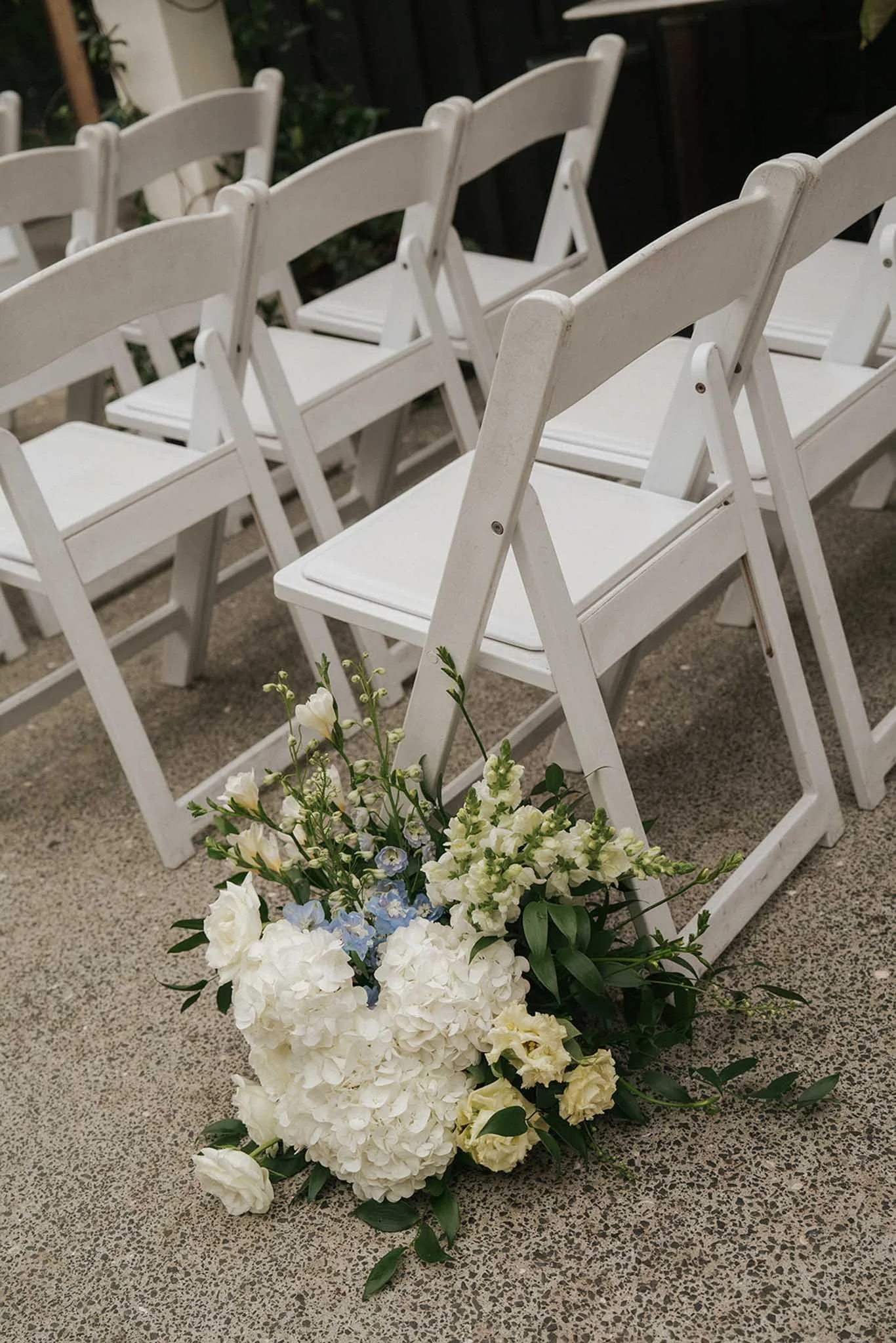 White folding chairs arranged in rows, with a floral arrangement of white and light blue flowers at the front, likely set up for an outdoor wedding or event.