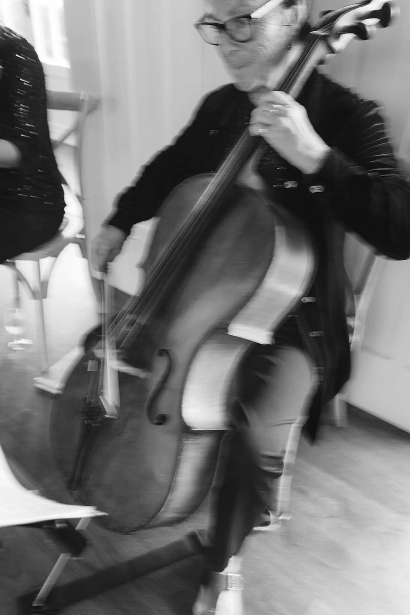 A woman with short hair, glasses, and a patterned shirt is playing a cello. She is sitting on a chair and focused on her music. There are chairs and a window in the background.