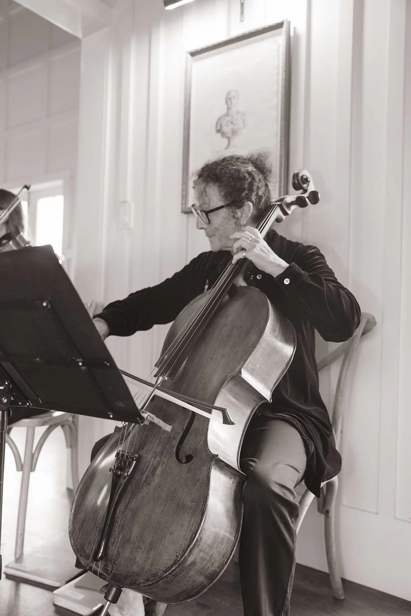 A woman playing a cello indoors, seated on a chair, reading sheet music, with white wooden panel walls and a framed portrait in the background.
