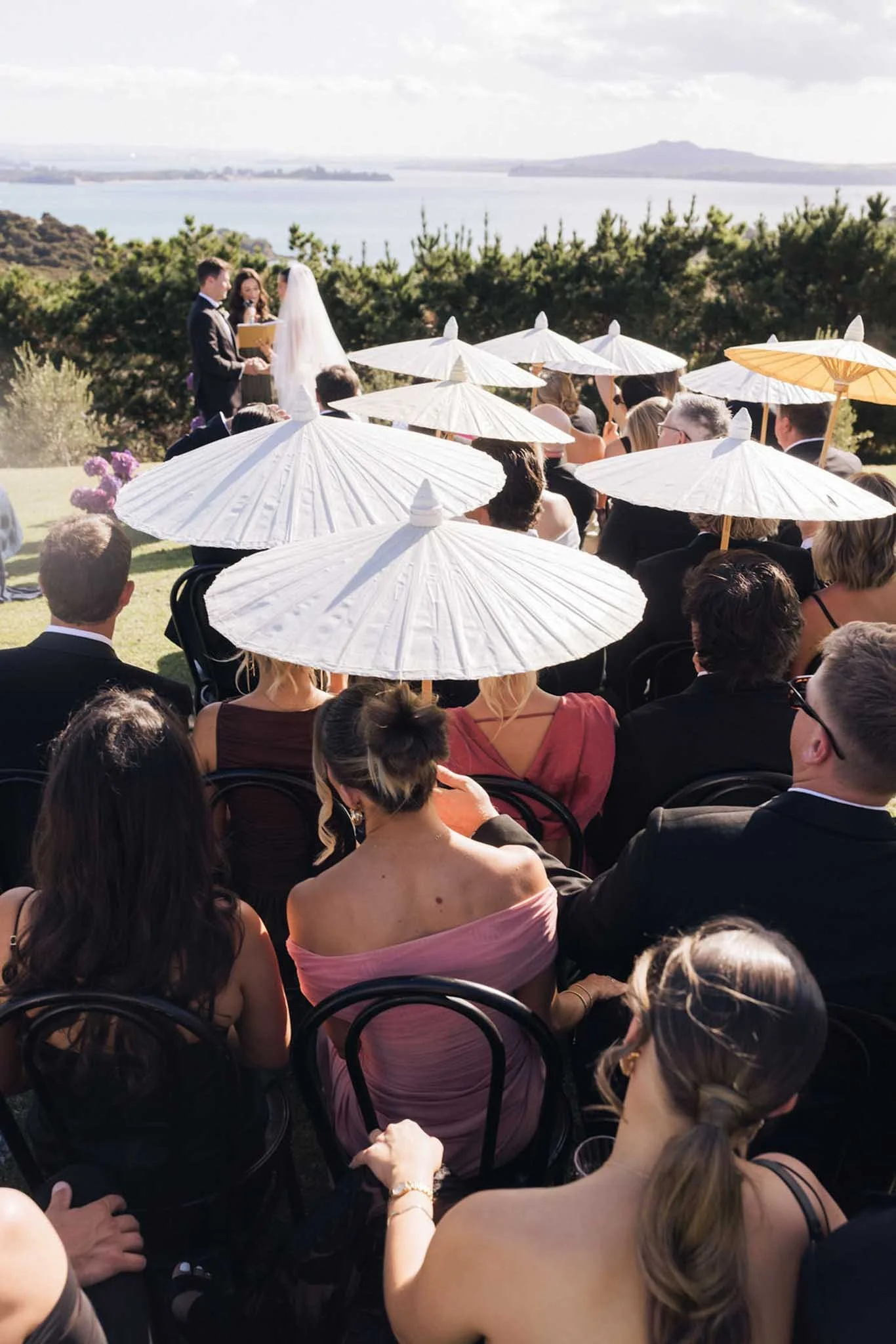 Outdoor wedding ceremony with guests sitting under white umbrellas overlooking a lake and mountains in the distance, with a couple exchanging vows at the altar.