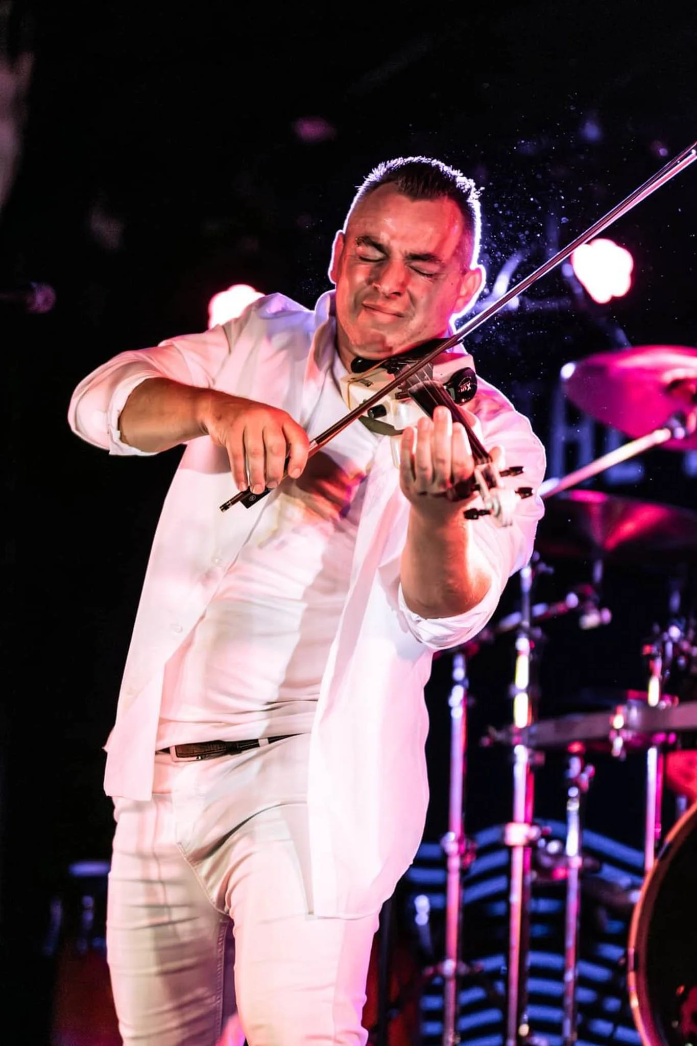 Man in white suit passionately playing the violin on stage with dark background and stage lighting
