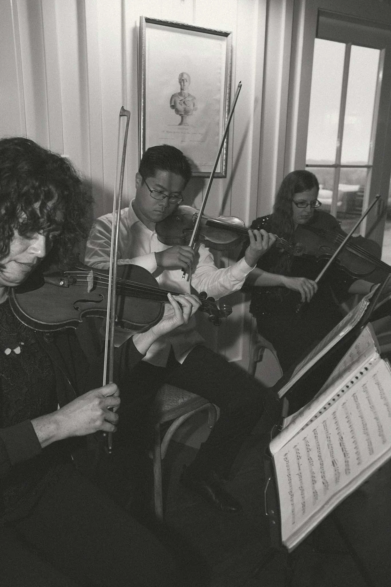 Three musicians playing string instruments, including violins and a viola, during a practice session in a room with a framed bust drawing on the wall and sheet music on a stand.