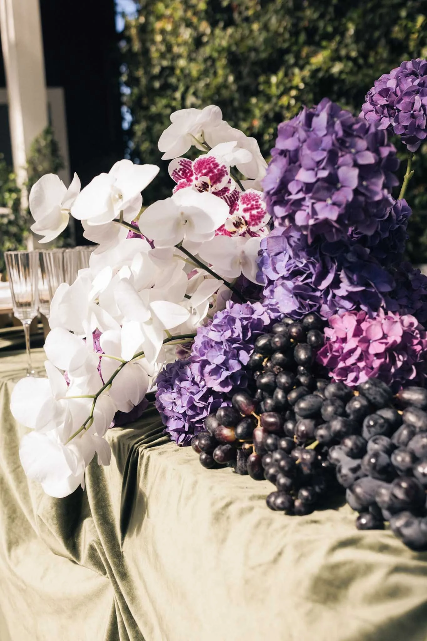Close-up of white orchids, purple hydrangeas, and black berries on a table covered with a beige cloth, set outdoors with trees and a building in the background.