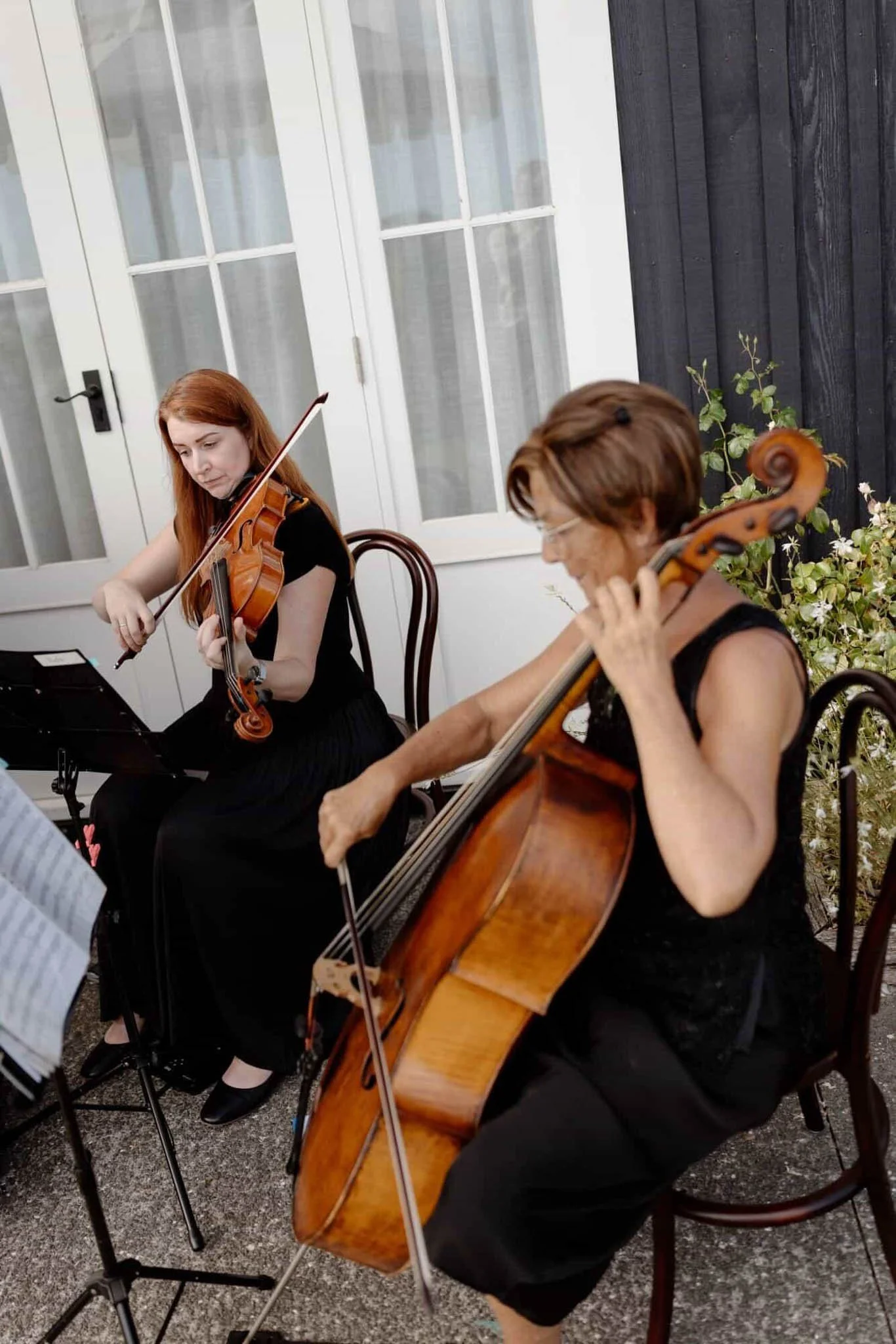 Two women playing musical instruments, one on the violin and the other on the cello, during a performance or rehearsal outside near a white door with glass panels, black siding, and plants.