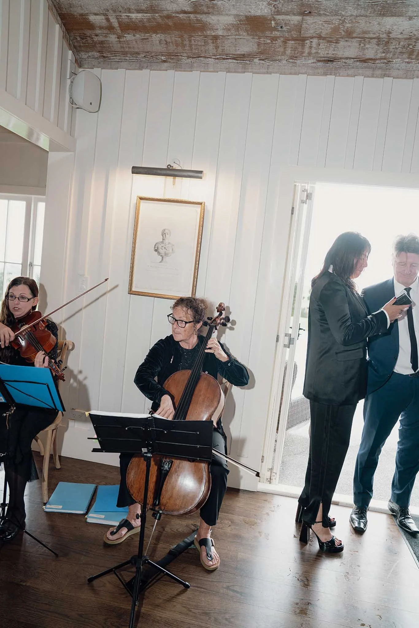 A group of musicians performing indoors, with some standing and others sitting, in a room with white panel walls and wooden flooring. There are two women with violins and one woman with a cello, and a man and woman standing near a door, possibly in c