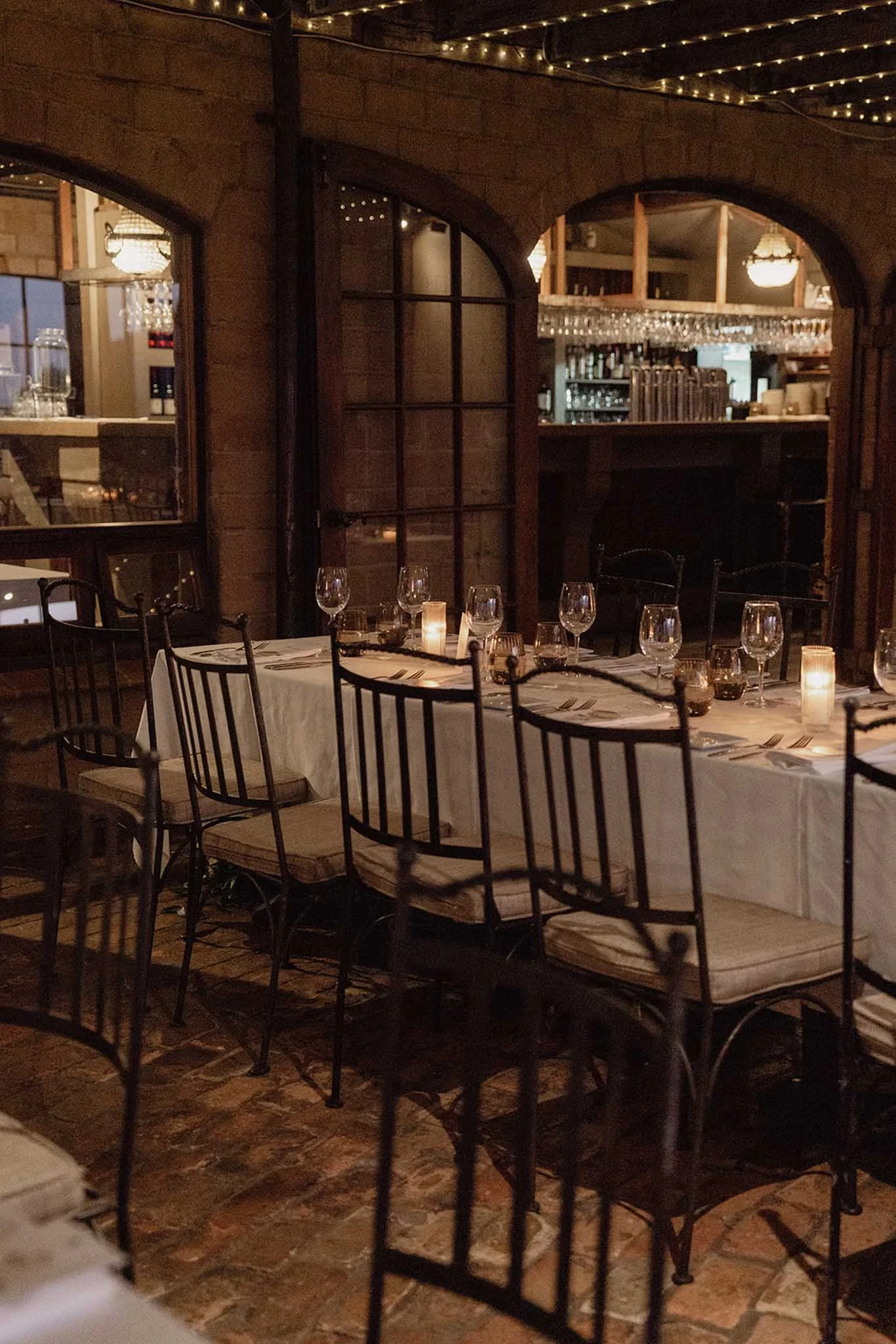 A restaurant interior with a long table set with multiple wine glasses, candles, and tableware. In the background, there are arched windows and a bar with hanging glasses and shelves of bottles. The ceiling is decorated with string lights, creating a