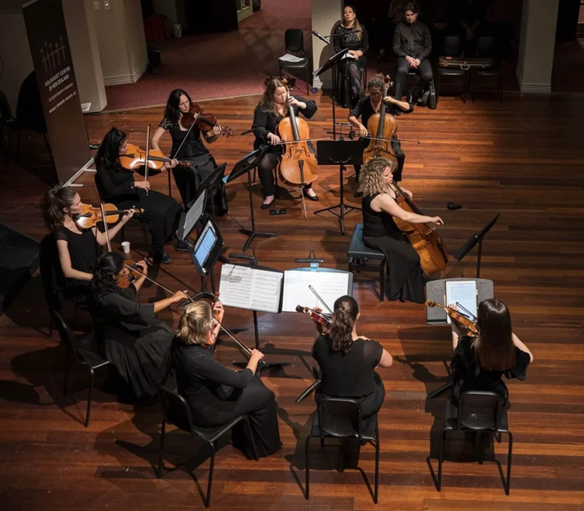 Orchestra with musicians playing violins and cellos on a wooden stage, with music stands and sheet music in an indoor concert hall.