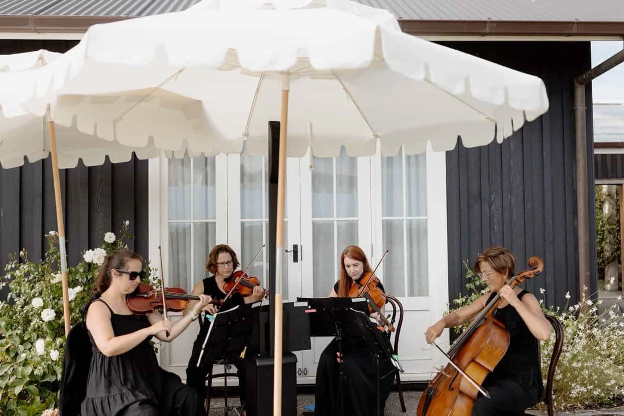 Group of women playing string instruments outdoors under white patio umbrellas, with black building and windows in background.