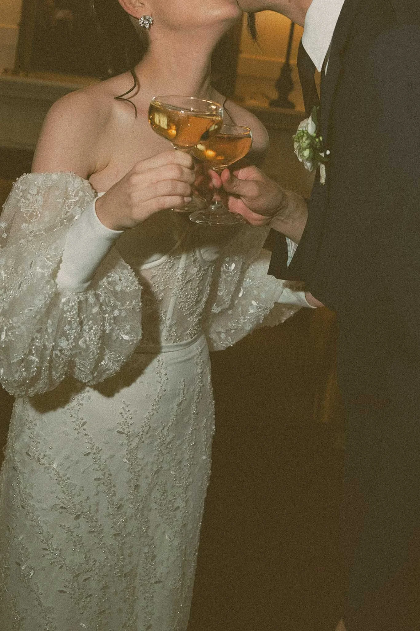 A bride and groom are holding champagne glasses in a toast during their wedding. The bride wears a lace wedding dress and elaborate earrings. The groom wears a dark suit with a boutonniere. They are close together, sharing a kiss.