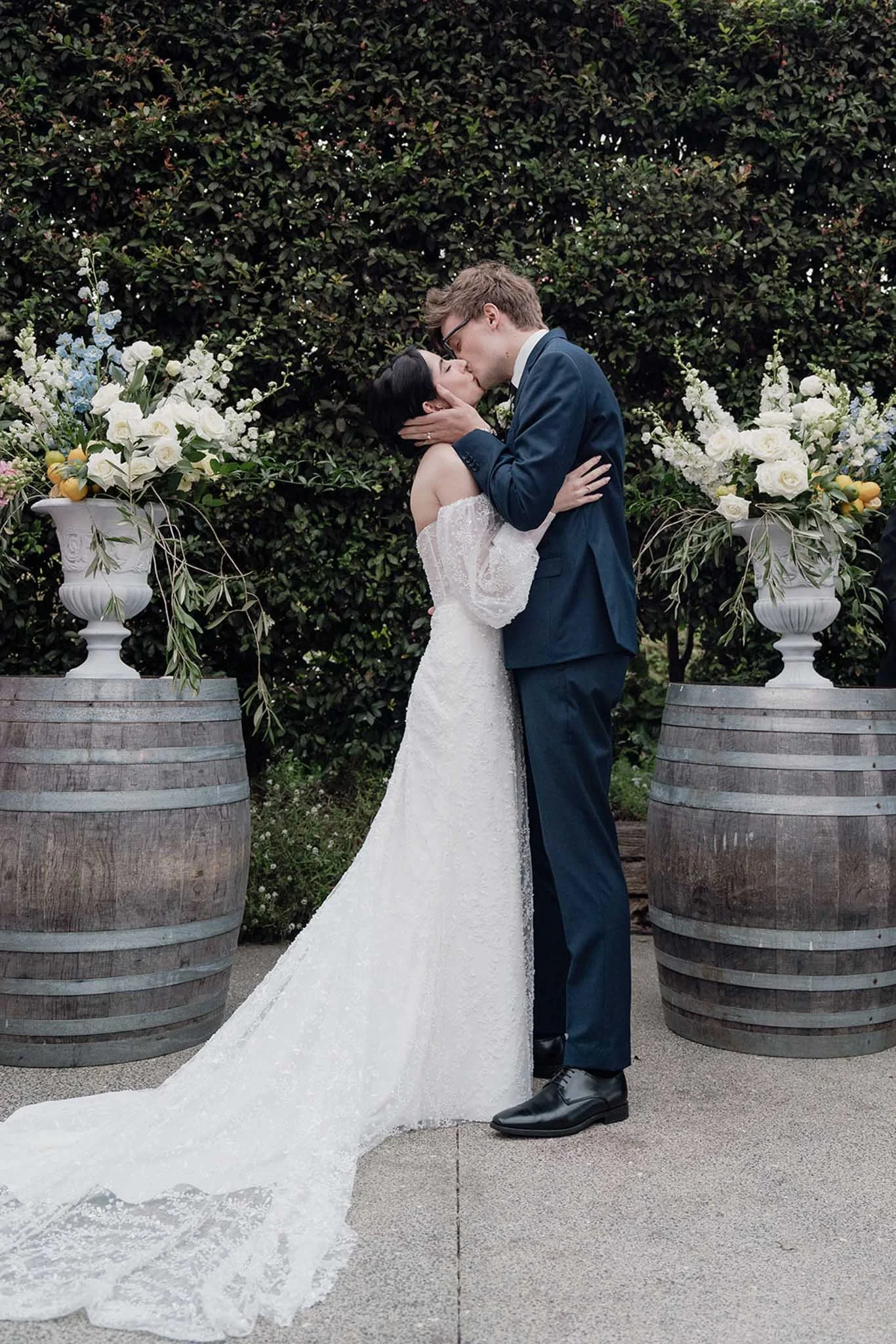 A bride and groom share a kiss, with roses, white lilies, and greenery in large white urns flanking them, against a dense green hedge.