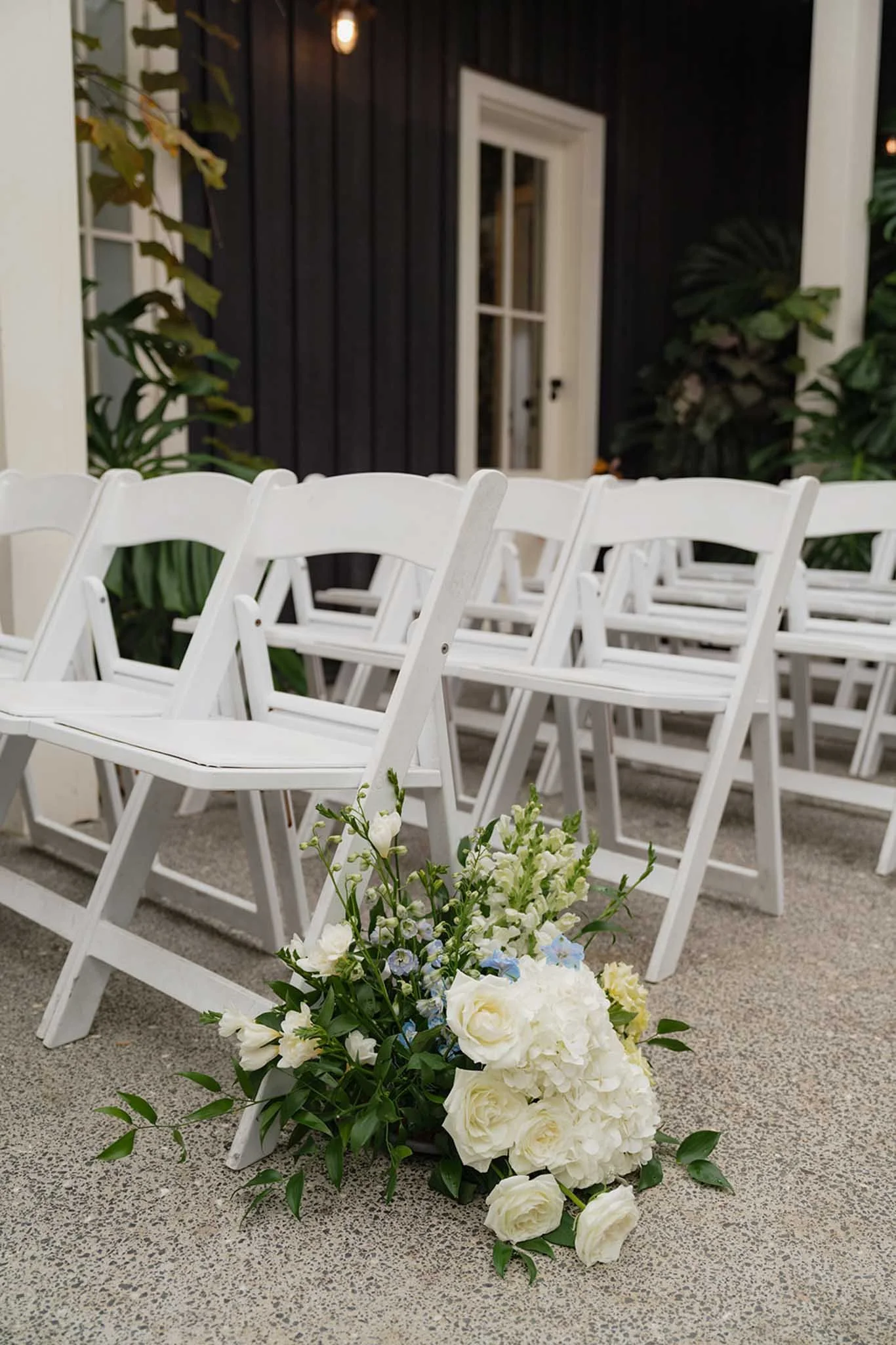 White chairs arranged outdoors with a floral centerpiece in front, against a backdrop of a black building with white trim and a door, surrounded by greenery.