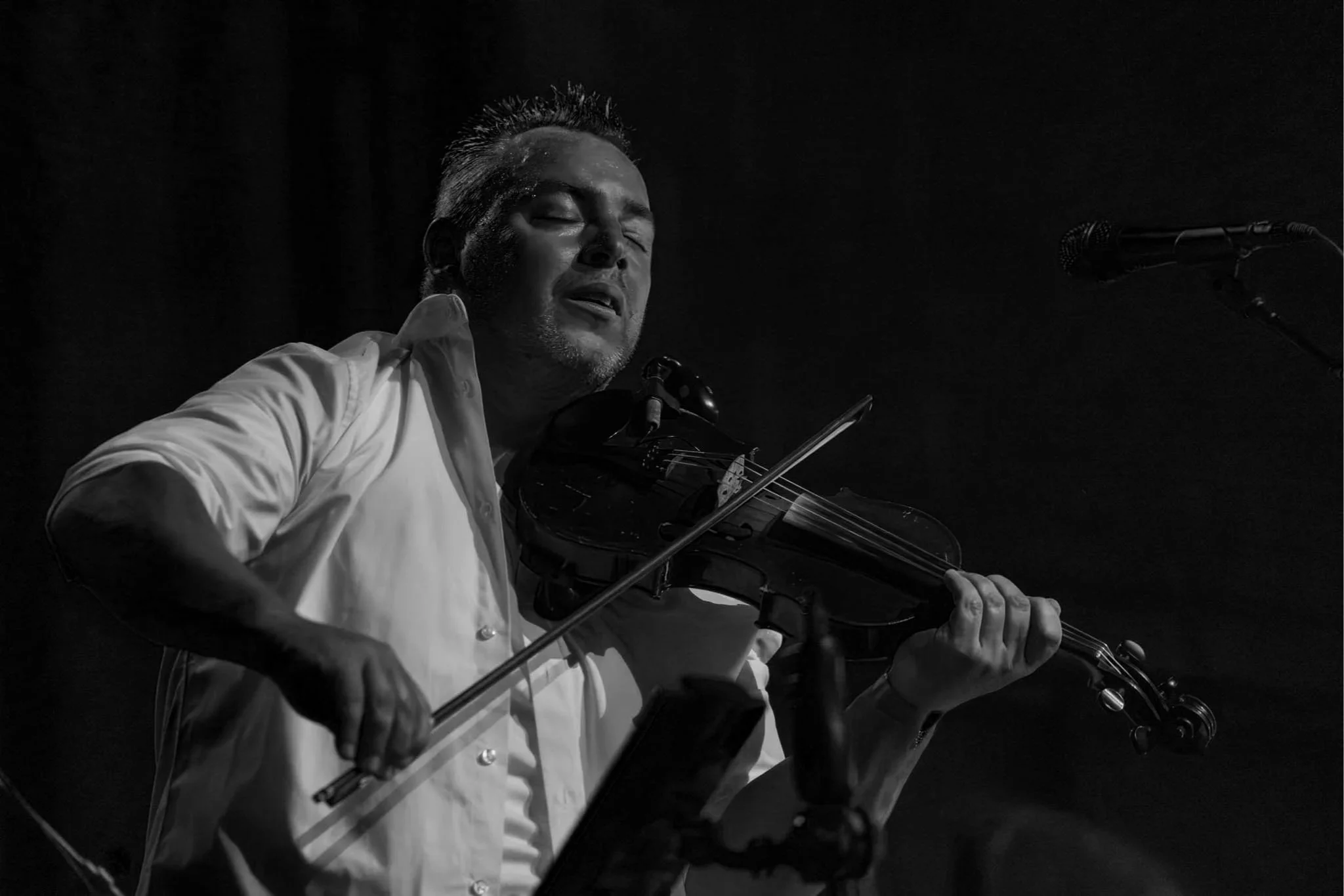A man playing the violin with closed eyes, wearing a white shirt, in a dark setting.
