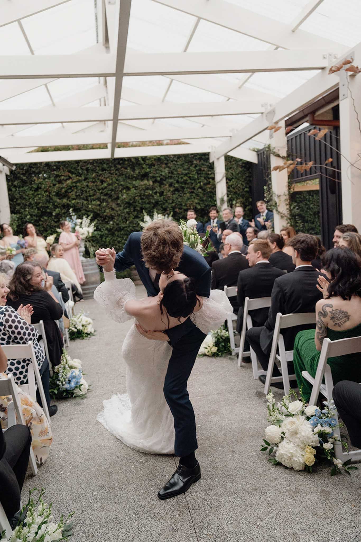 A wedding ceremony with a bride and groom dancing in front of seated guests under a white canopy with green foliage and flowers.