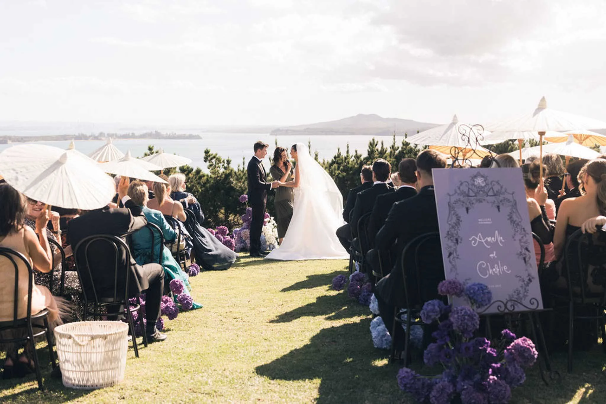 Outdoor wedding ceremony with a lake and mountains in the background. Bride and groom holding hands at the altar. Guests seated under umbrellas, decorated with purple flowers.