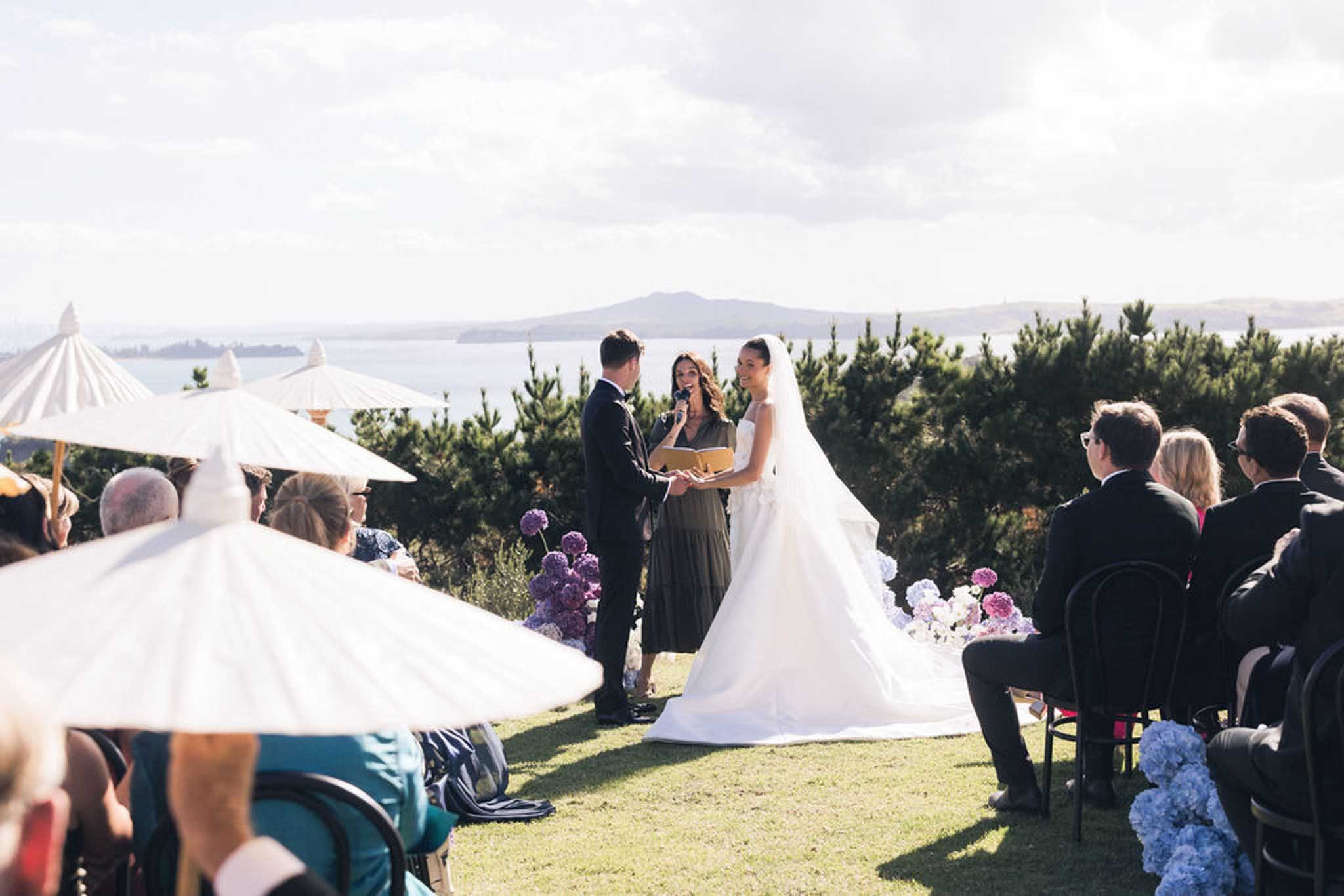 A wedding ceremony outdoors with the bride and groom exchanging vows, surrounded by guests seated under umbrellas with scenic water and mountains in the background.