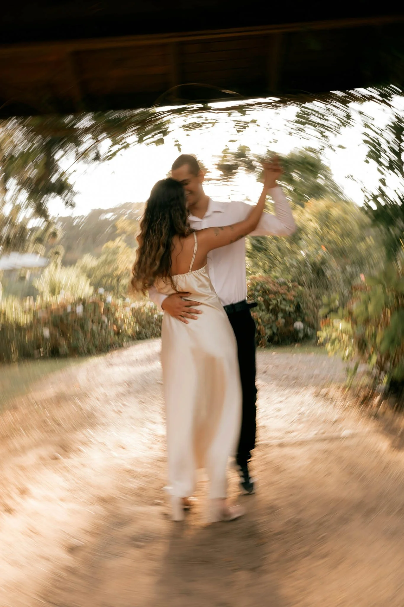 A couple dancing outdoors during sunset, the woman has long wavy hair and is wearing a cream dress, the man is in a white shirt and dark pants, and they are embracing each other.