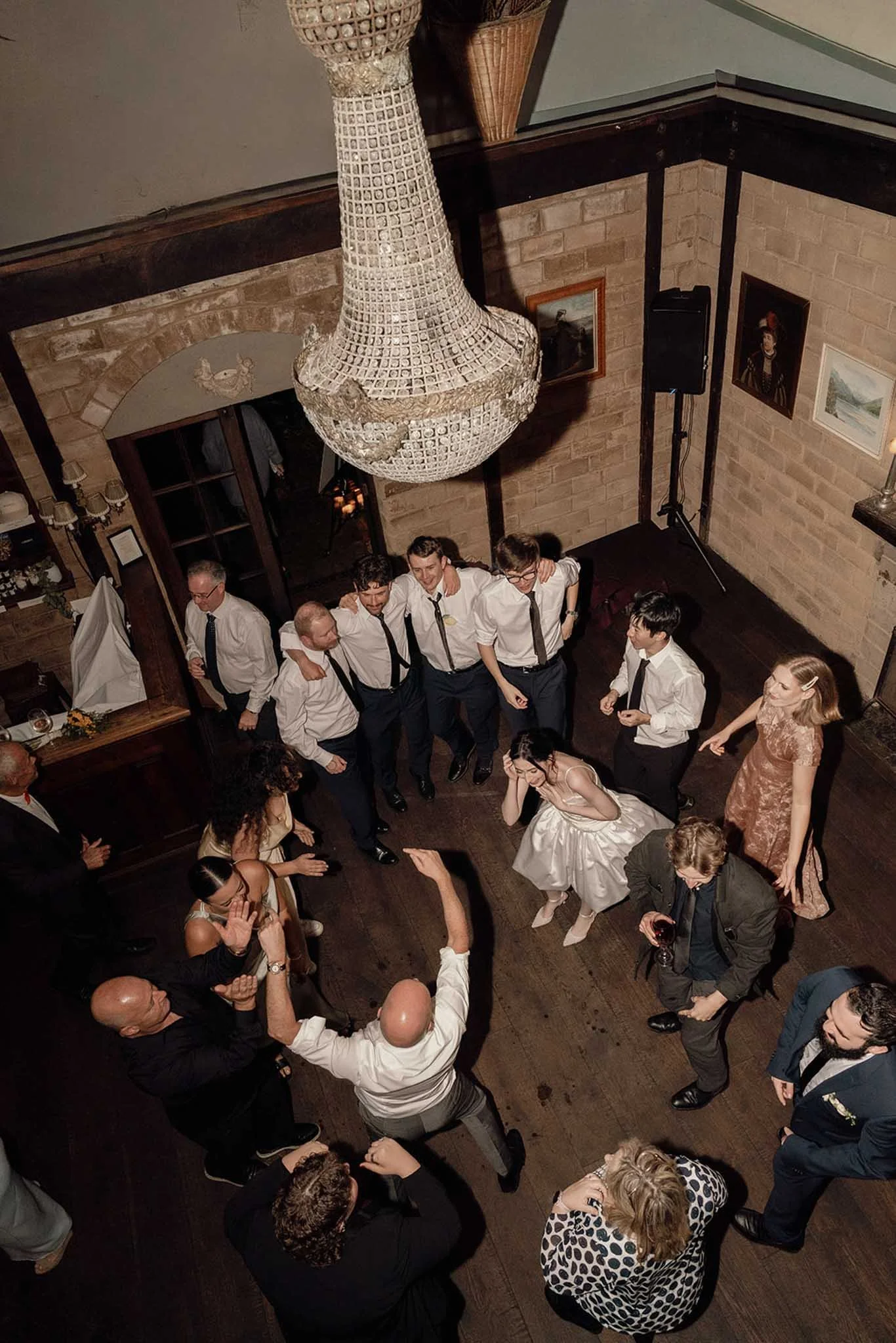 People dancing and socializing in a warmly lit venue with a large, ornate chandelier hanging from the ceiling.