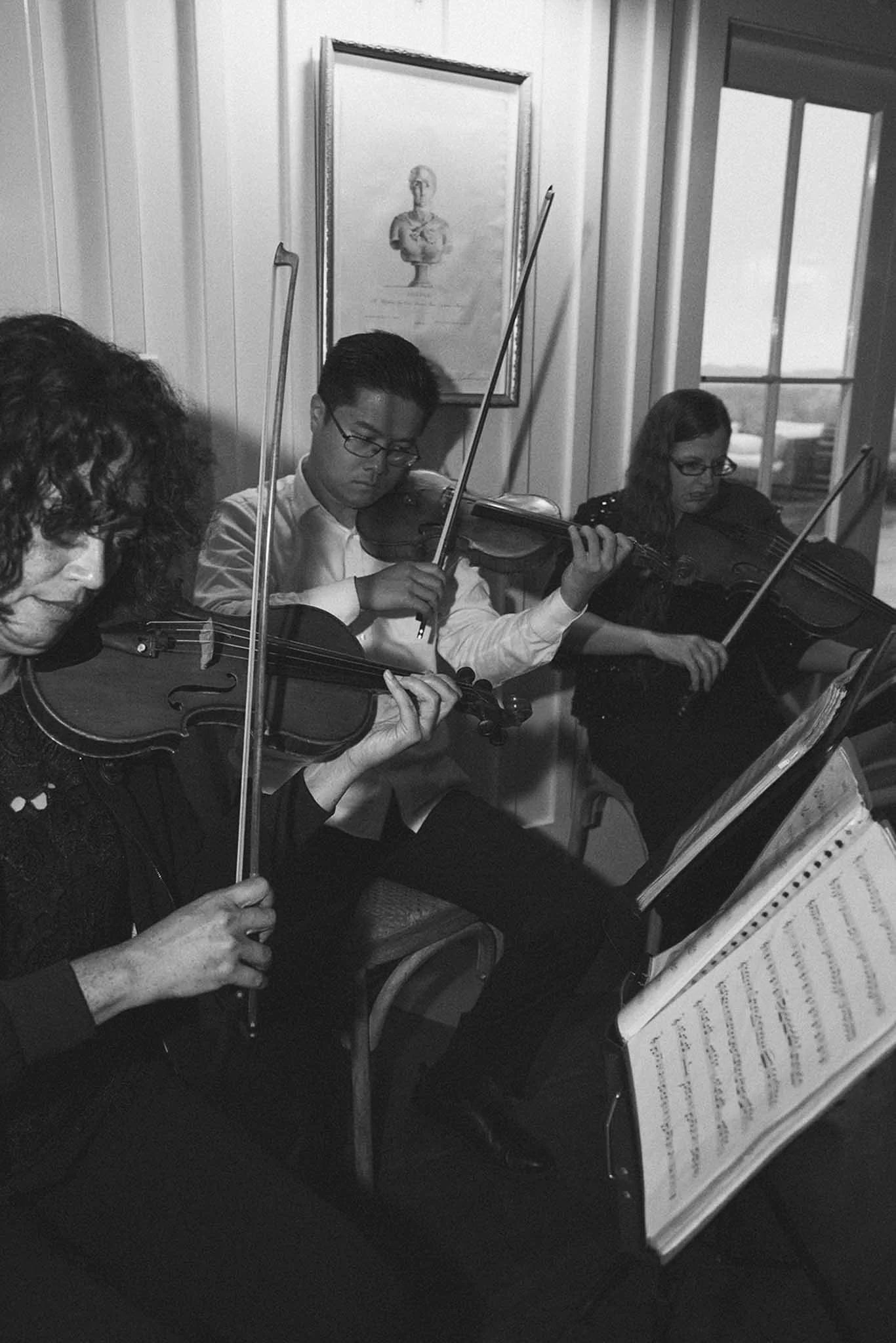 Black and white photo of three musicians playing violins during a practice or performance, with sheet music on a stand in front of them, in a room with a framed picture on the wall and windows showing an outdoor scene.