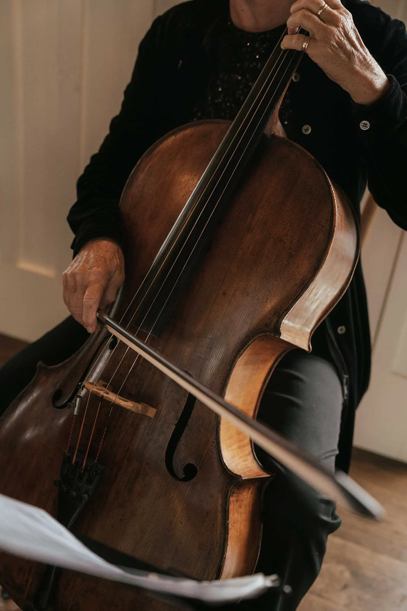 Close-up of a person playing a wooden cello, showing their hand on the neck of the instrument and part of their face and body. The person is wearing a black top with buttons and rings on their fingers.