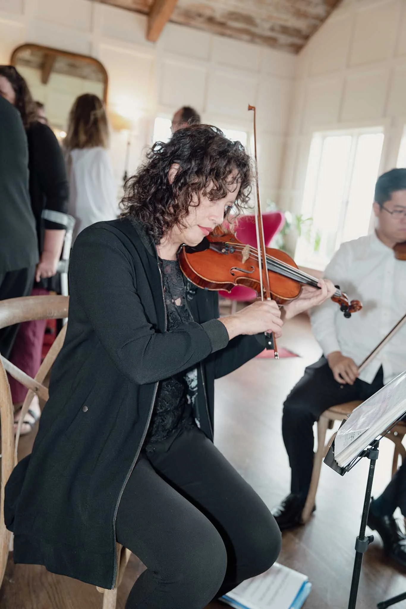 A woman with curly dark hair playing the violin while sitting on a chair, with other people and music stands around her in a cozy room with natural light.