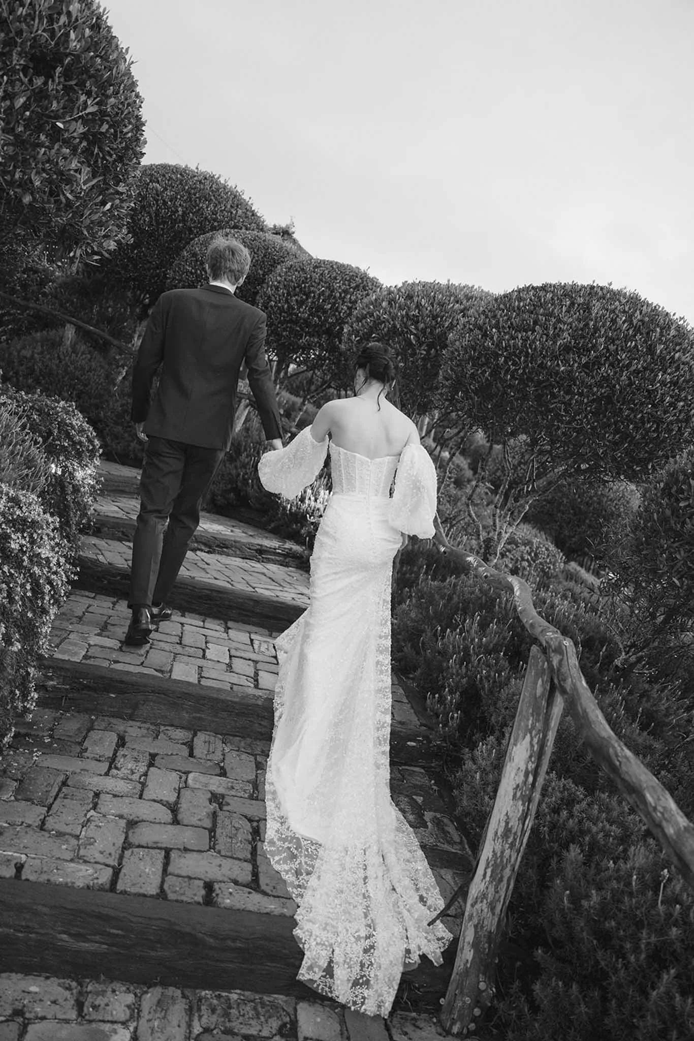 A man in a suit holding hands with a woman in a wedding dress walking along a garden path with trees and bushes.