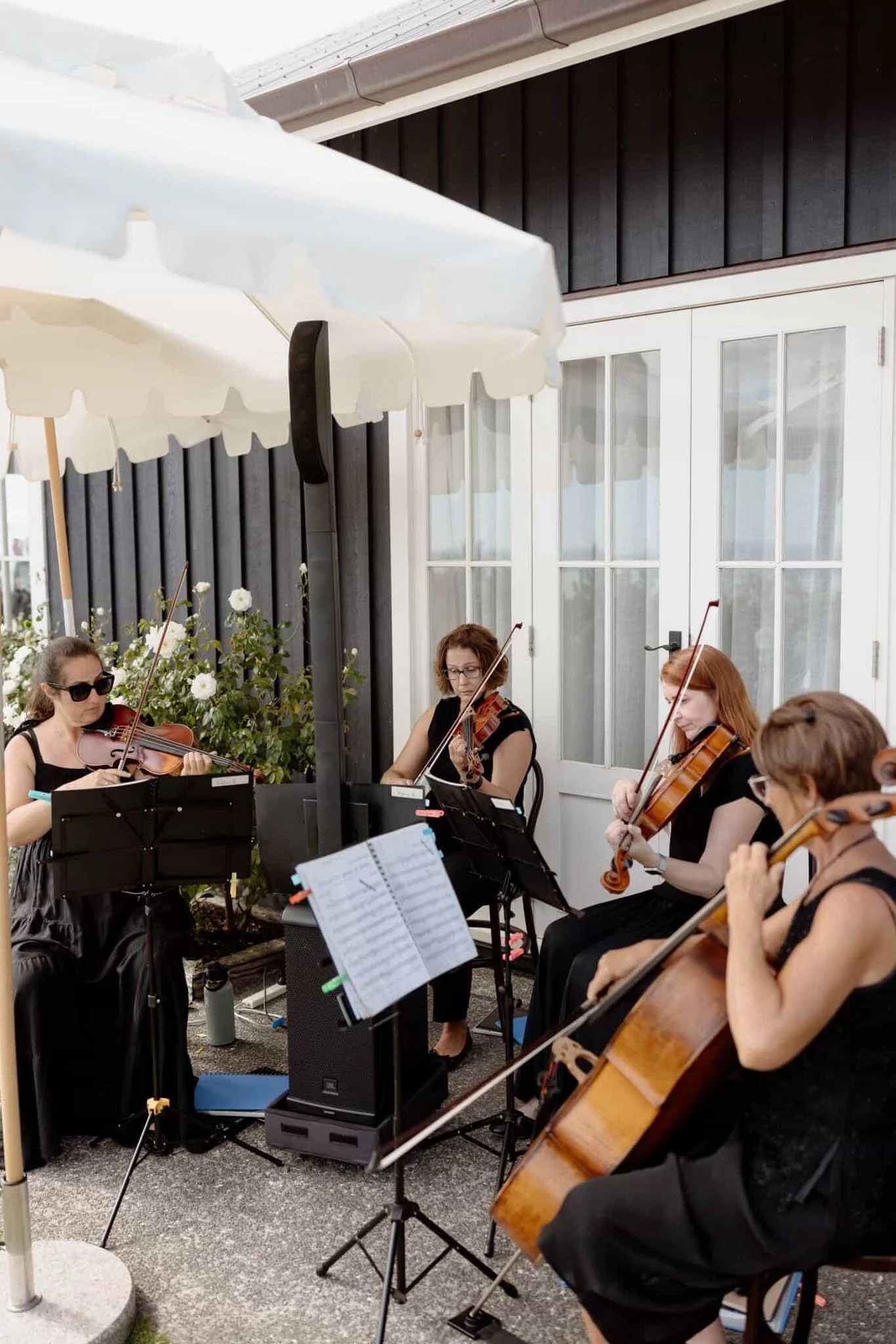 A group of women playing string instruments, such as violins and a cello, outdoors under a band of white patio umbrellas, near a building with white doors and black siding, in a garden setting.