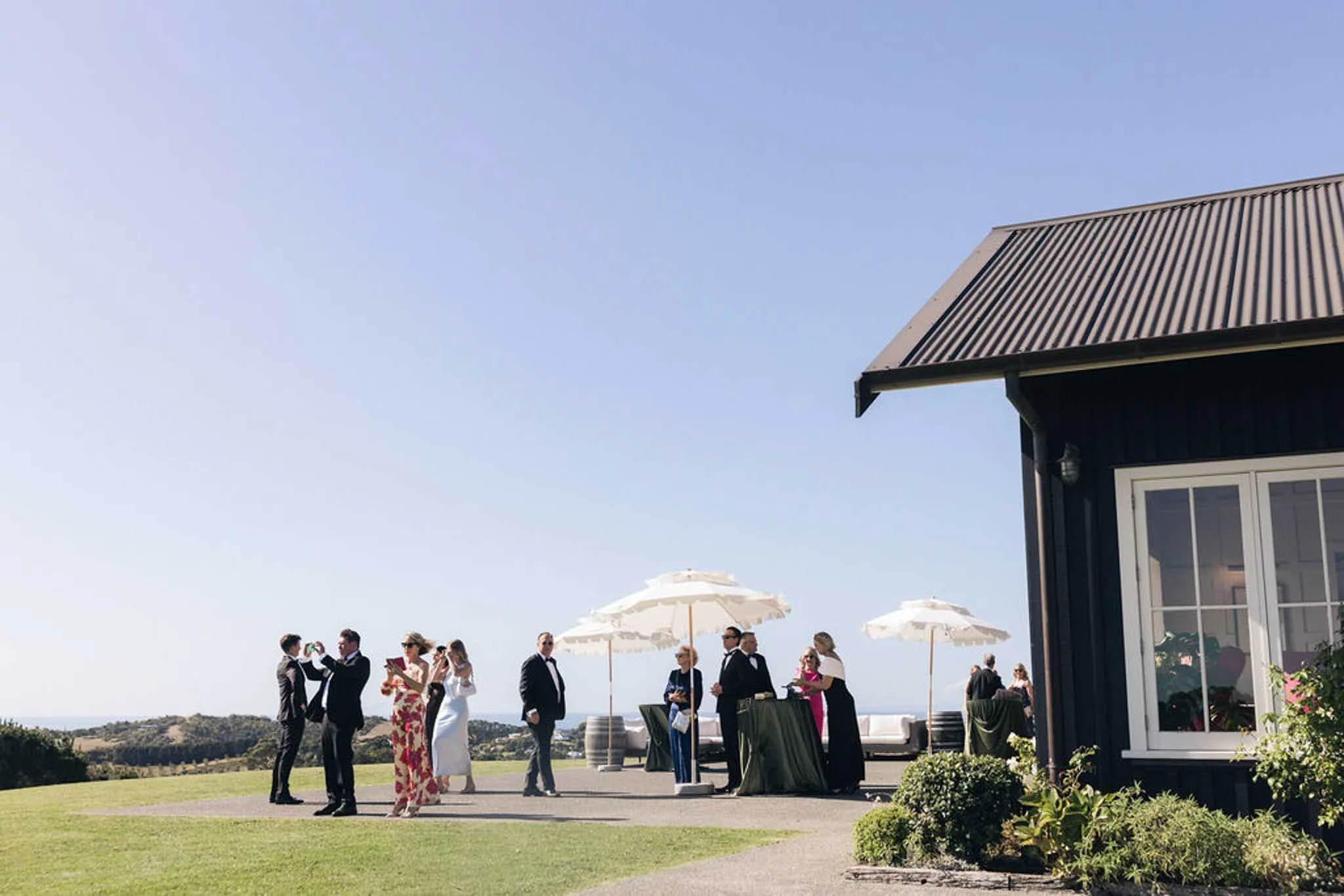 People dressed in formal attire attending an outdoor event under umbrellas on a sunny day, with a building featuring a gabled roof and a view of distant hills.