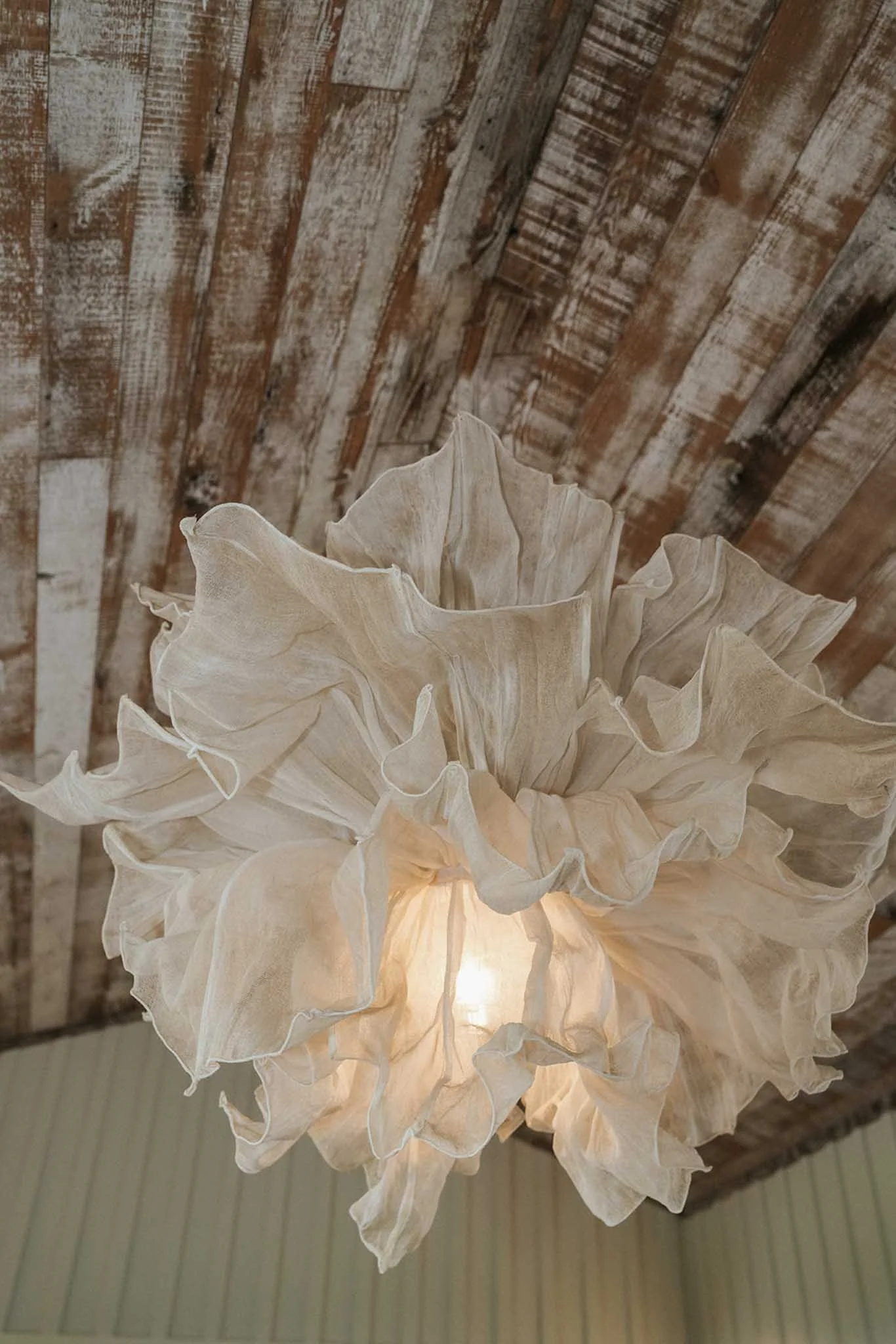 A close-up of a floral chandelier made of cream-colored fabric with a wooden ceiling background.