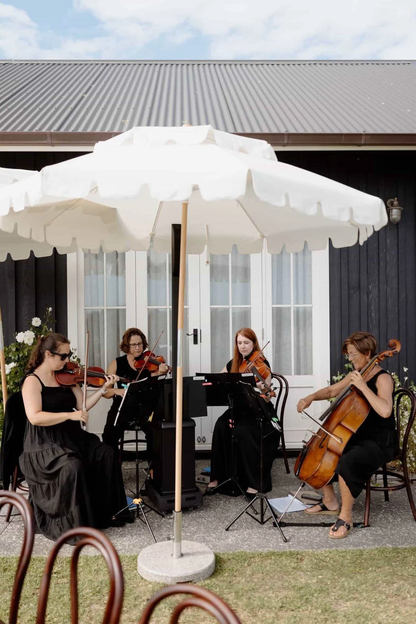 A group of women playing string instruments such as violins and a cello in an outdoor setting, under a large white umbrella, with a dark building and a blue sky with clouds in the background.