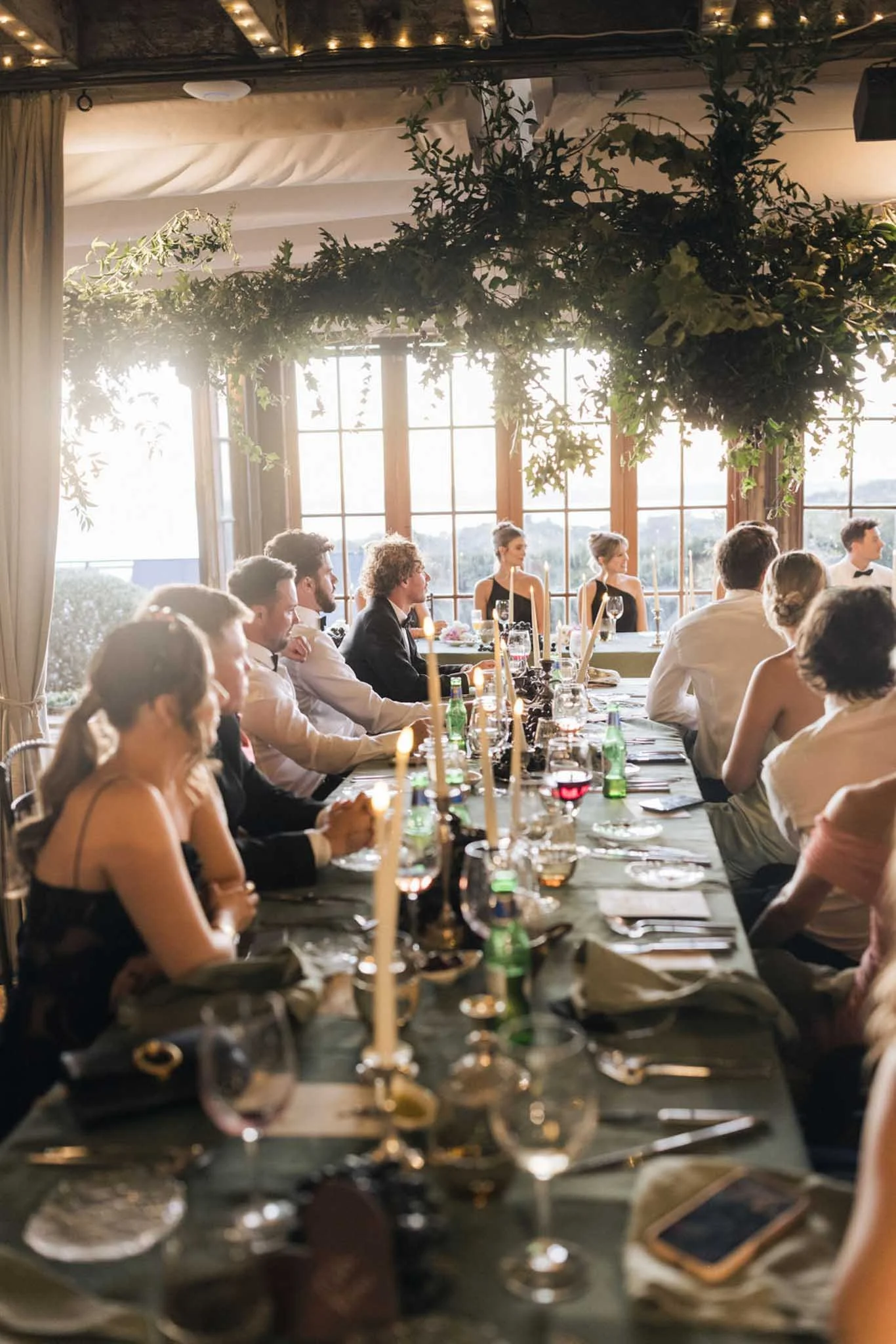 People sitting at a long banquet table in a warmly-lit indoor venue with large windows and greenery hanging from the ceiling, celebrating a formal event.