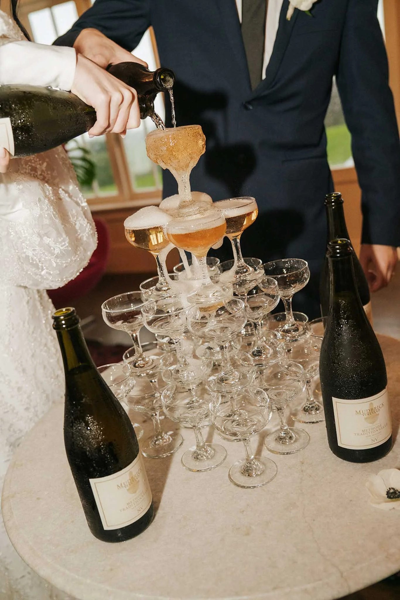 A champagne tower at a wedding reception with three bottles of champagne, a bride and groom pouring champagne into the top glass, and glasses arranged in a pyramid.