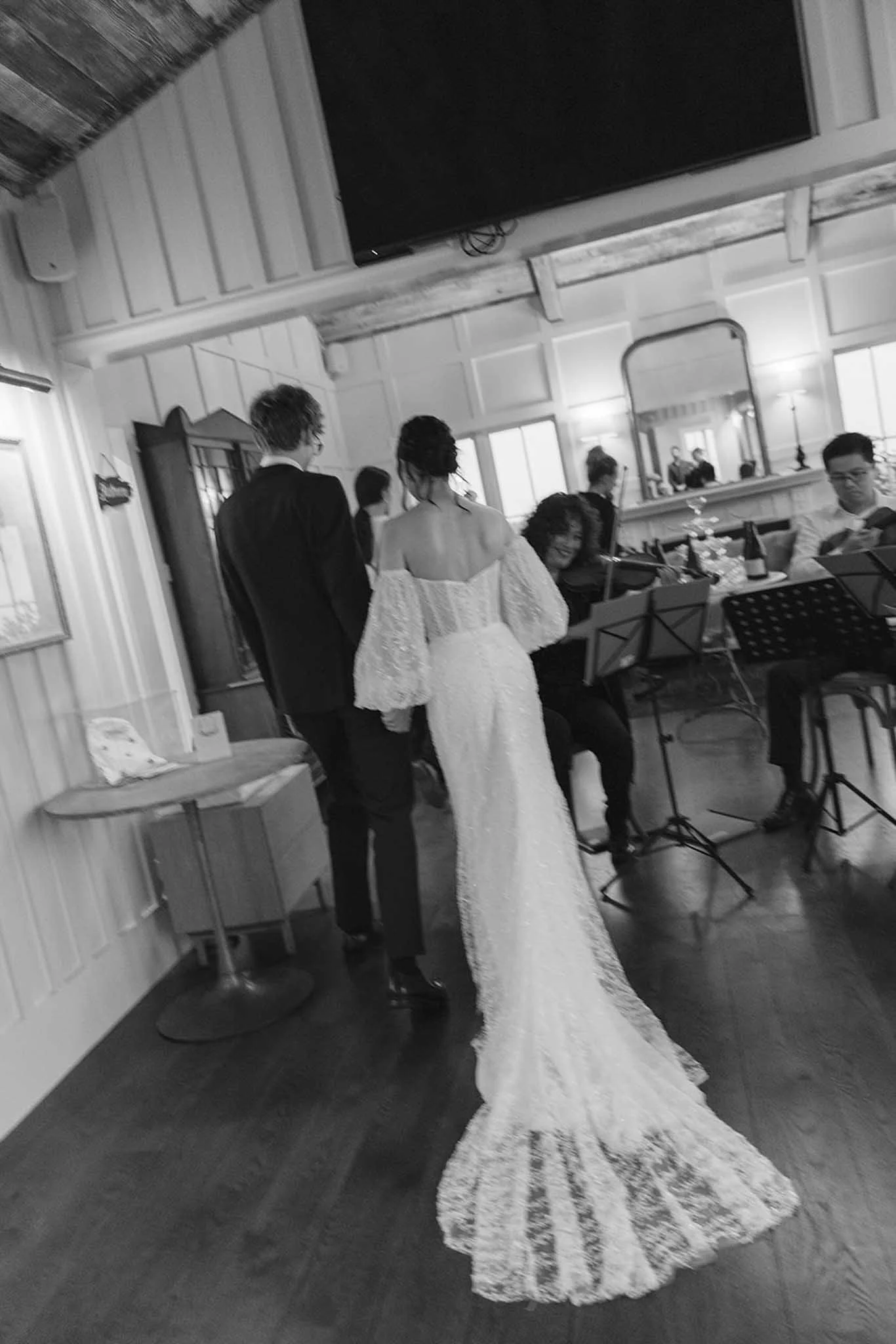 A couple dressed in wedding attire, the bride in a white gown with a long train and the groom in a black suit, holding hands and walking in a rustic indoor venue with wooden accents and a mirror.