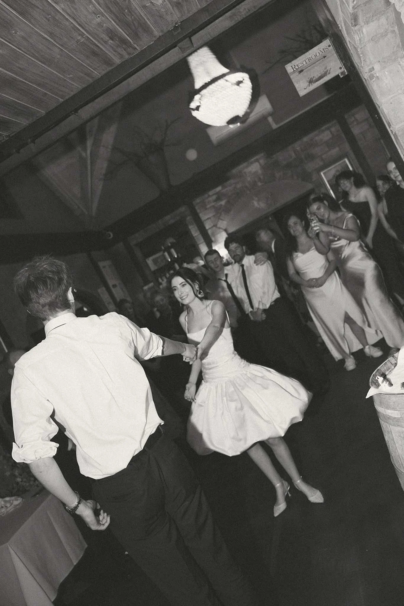A couple dancing at a wedding reception, holding hands and smiling. The bride is in a wedding gown, and the groom is in a dress shirt and pants. Other guests are seen in the background, and a chandelier hangs from the ceiling.
