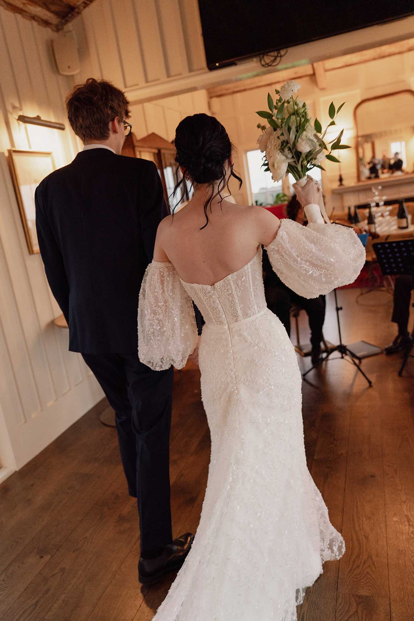 A bride in a white lace wedding dress holding a bouquet of white flowers and an officiant dressed in black suit standing next to her at a wedding ceremony indoors with wooden walls and a TV in the background.
