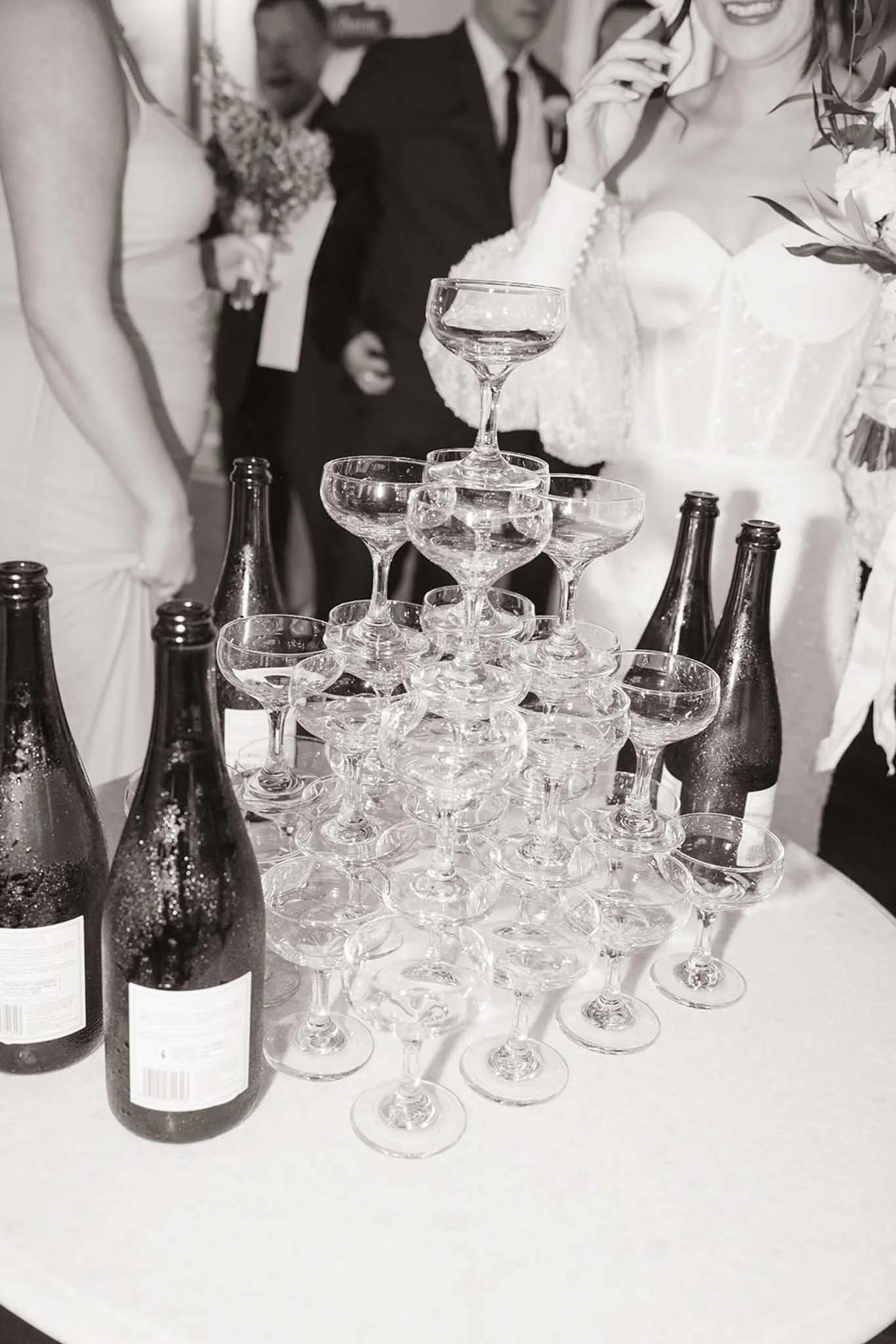 A table with multiple empty champagne glasses stacked in a pyramid shape, surrounded by several beer bottles, at a celebration or party with people in the background.