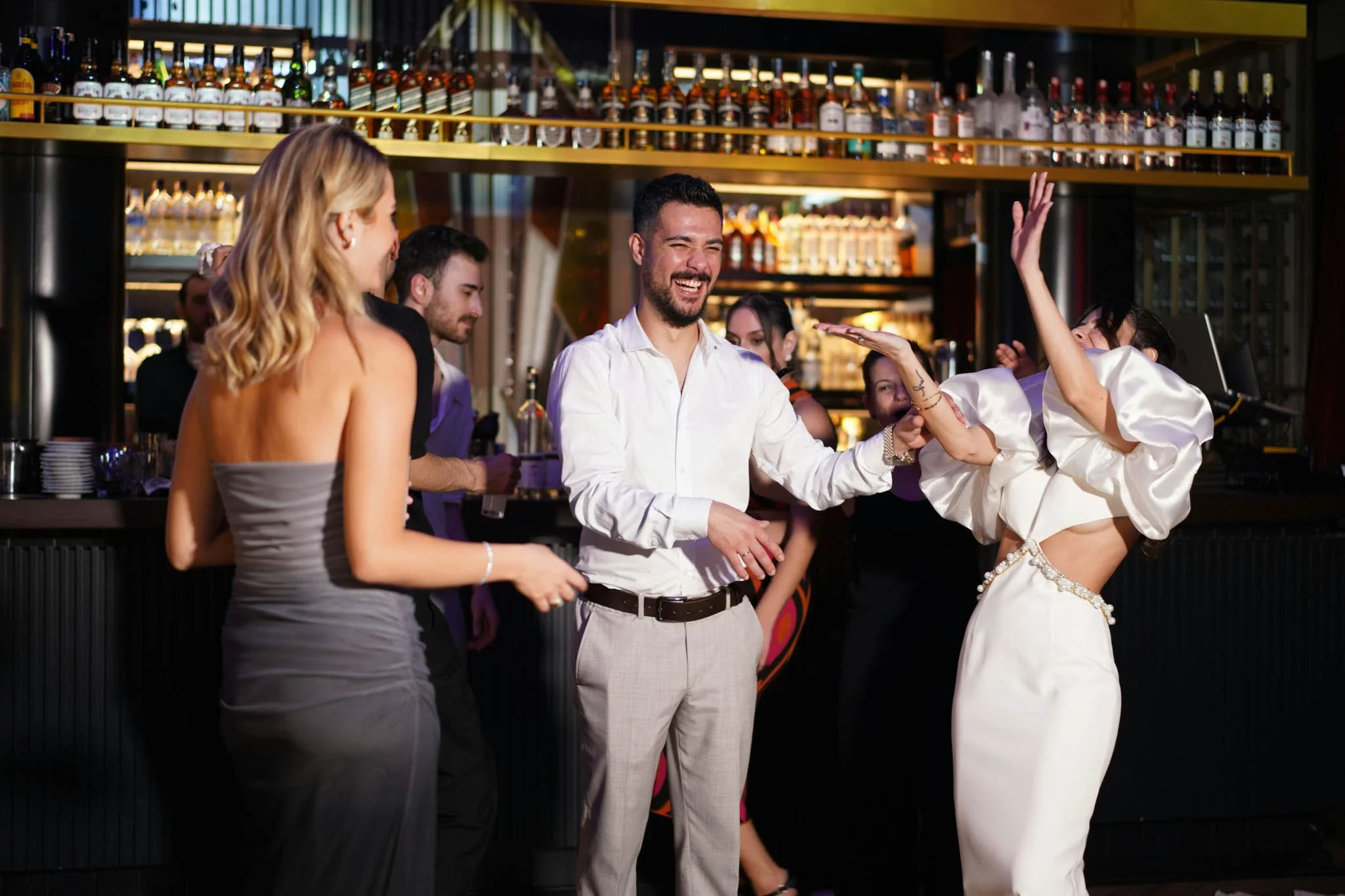 Group of people dancing and socializing at a bar or nightclub with bottles of alcohol on shelves in the background.