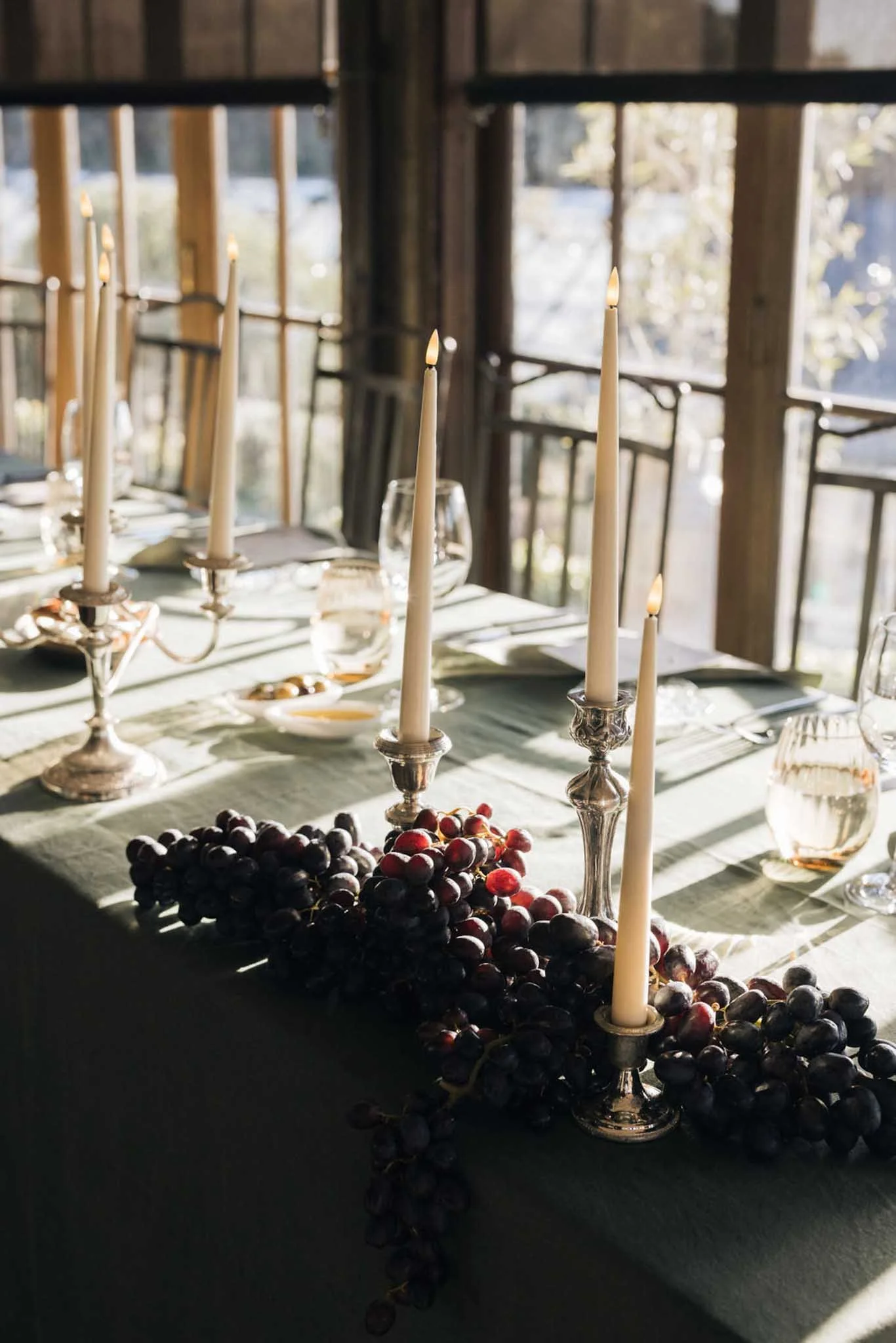 A table set for a meal with tall white candles in silver candle holders, glassware, and bunches of red and black grapes, in front of large windows with wooden frames letting in natural light.