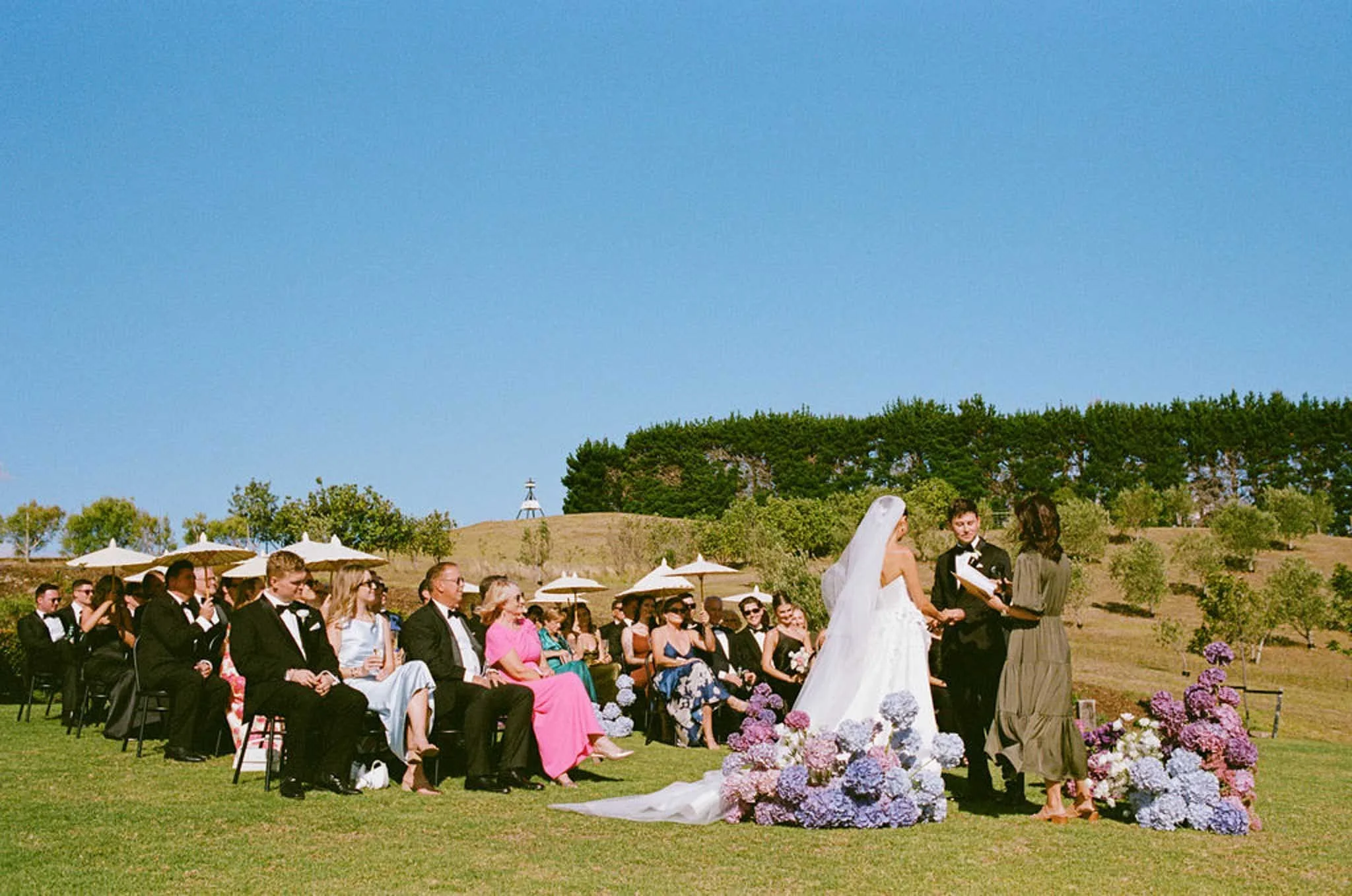 Outdoor wedding ceremony with seated guests in formal attire, officiant, bride, and groom exchanging vows, lush green landscape, blue sky, and floral arrangements at the altar.