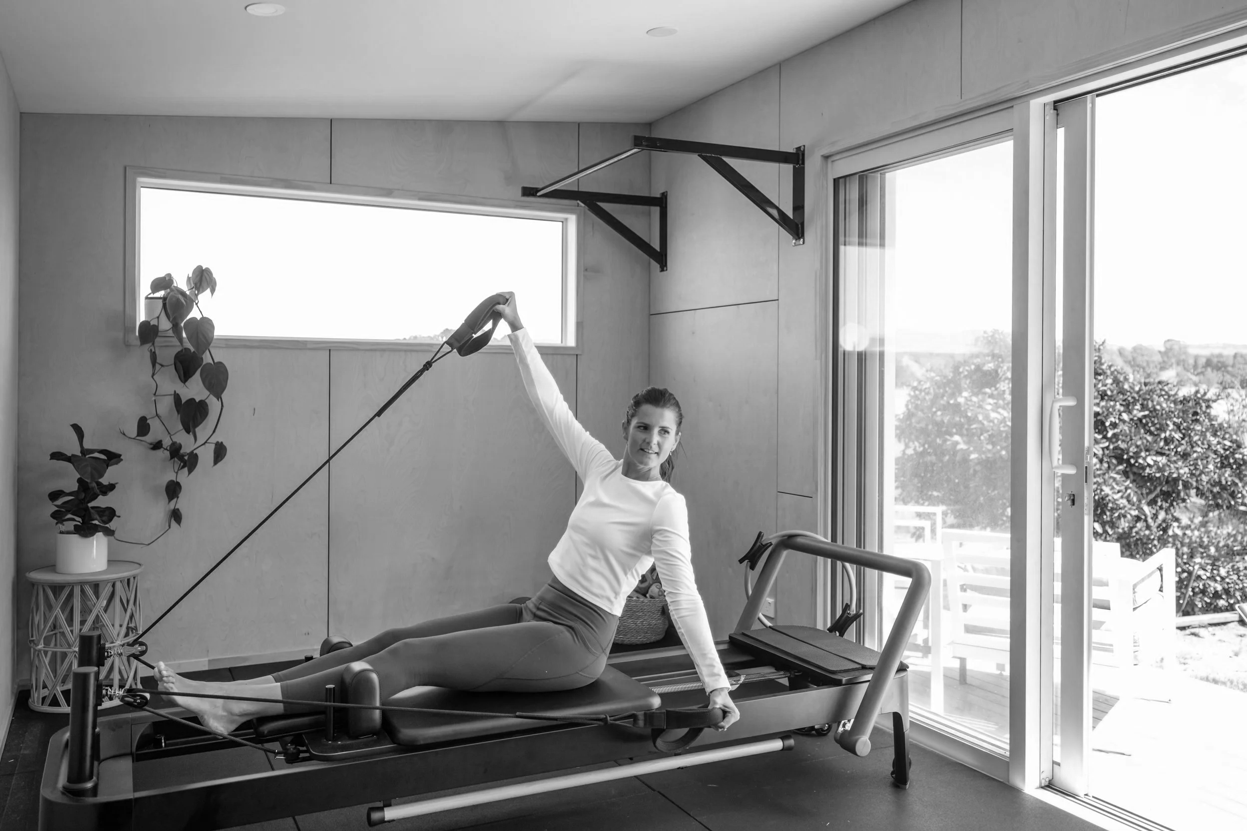 A woman performing a Pilates exercise on a reformer machine in a bright, modern fitness studio with large windows and a potted plant.