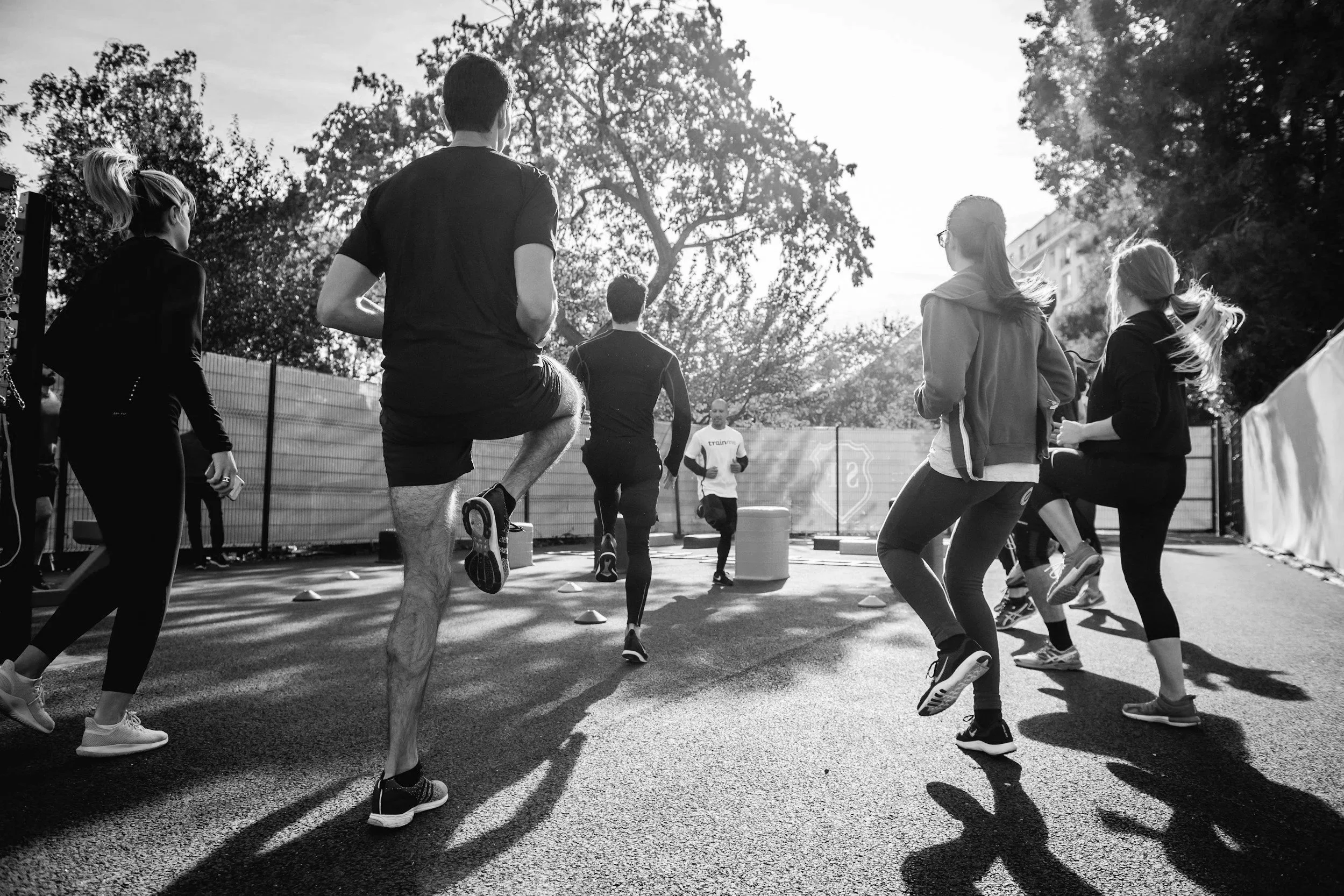 A group of people participating in an outdoor fitness class, running or jogging together on a paved surface with cones, surrounded by trees and a fence, with the sun shining brightly overhead.