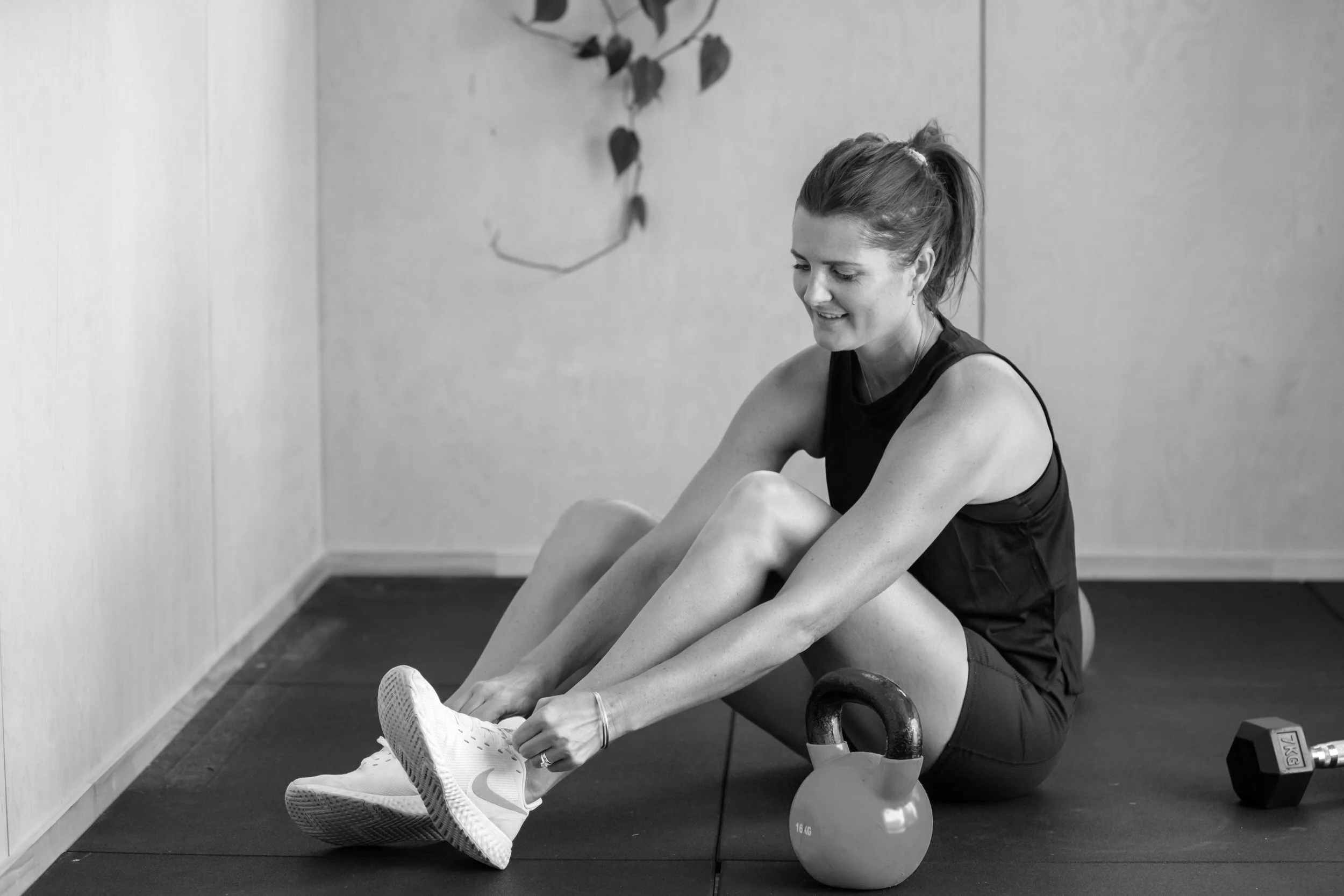 A woman with a ponytail in athletic clothing sits on a bench, tying her shoelace, with a kettlebell and a dumbbell nearby in a room with plain walls and a plant in the background.
