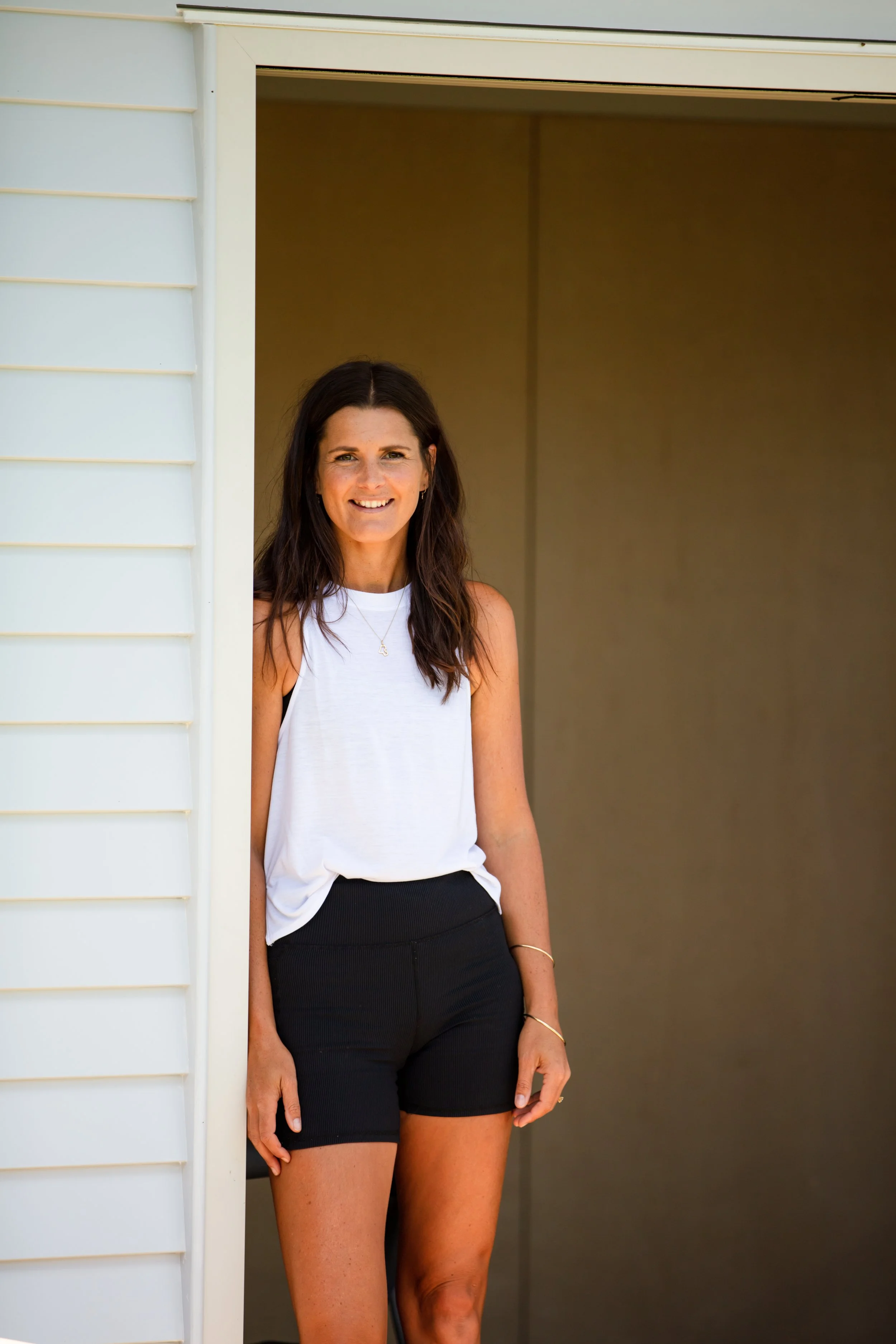 A woman with long dark hair smiling, wearing a white sleeveless top and black shorts, standing at a doorway outside the house.