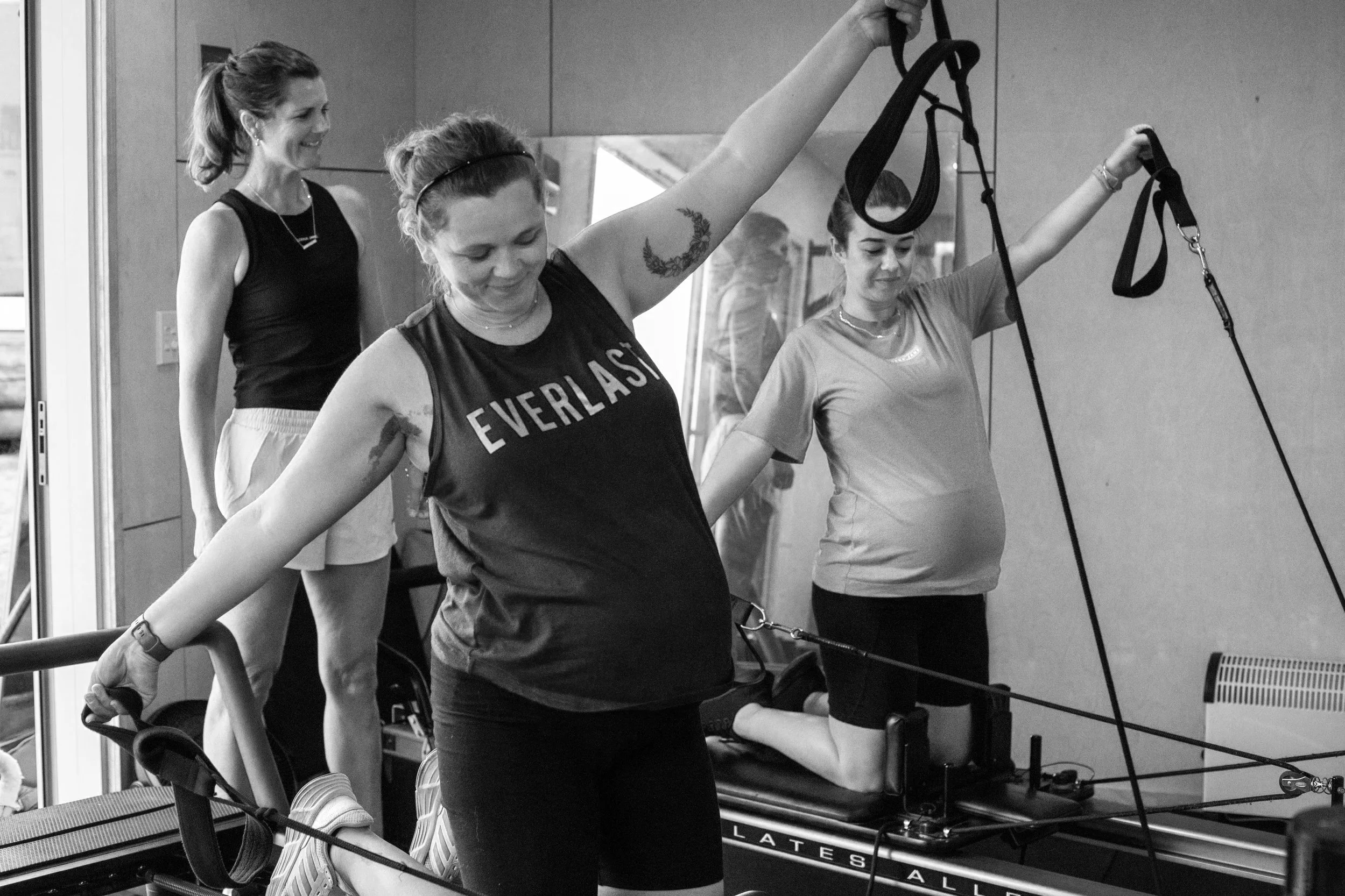 Three women exercising on a fitness reformer machine. One woman is standing, smiling, and watching the other two, who are lying face down on the machine. The women are in a gym or fitness studio with mirrors and equipment visible in the background.