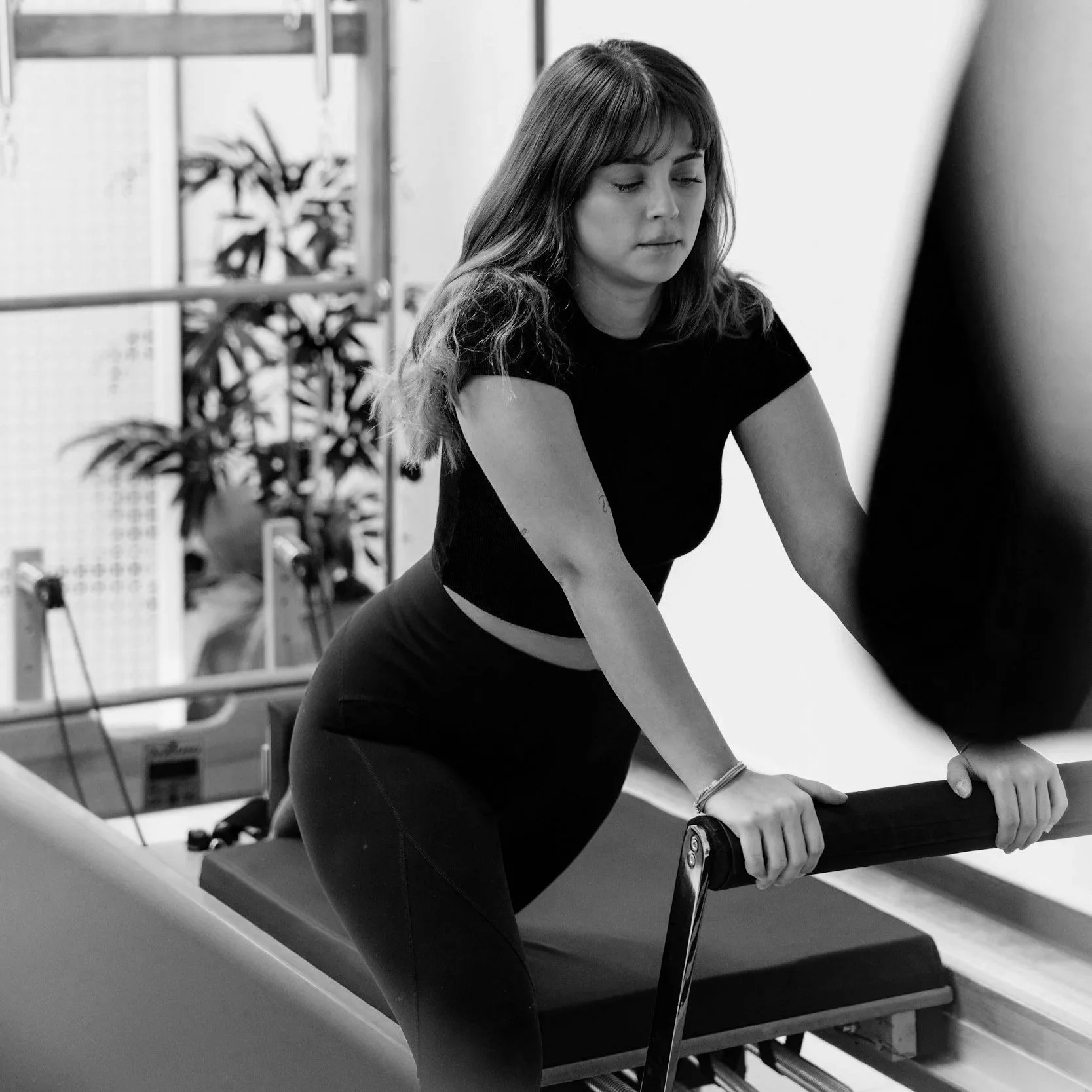 A woman with long hair wearing a black t-shirt and dark pants leaning on a pool table, holding its edge, in a room with indoor plants and pool cues in the background.