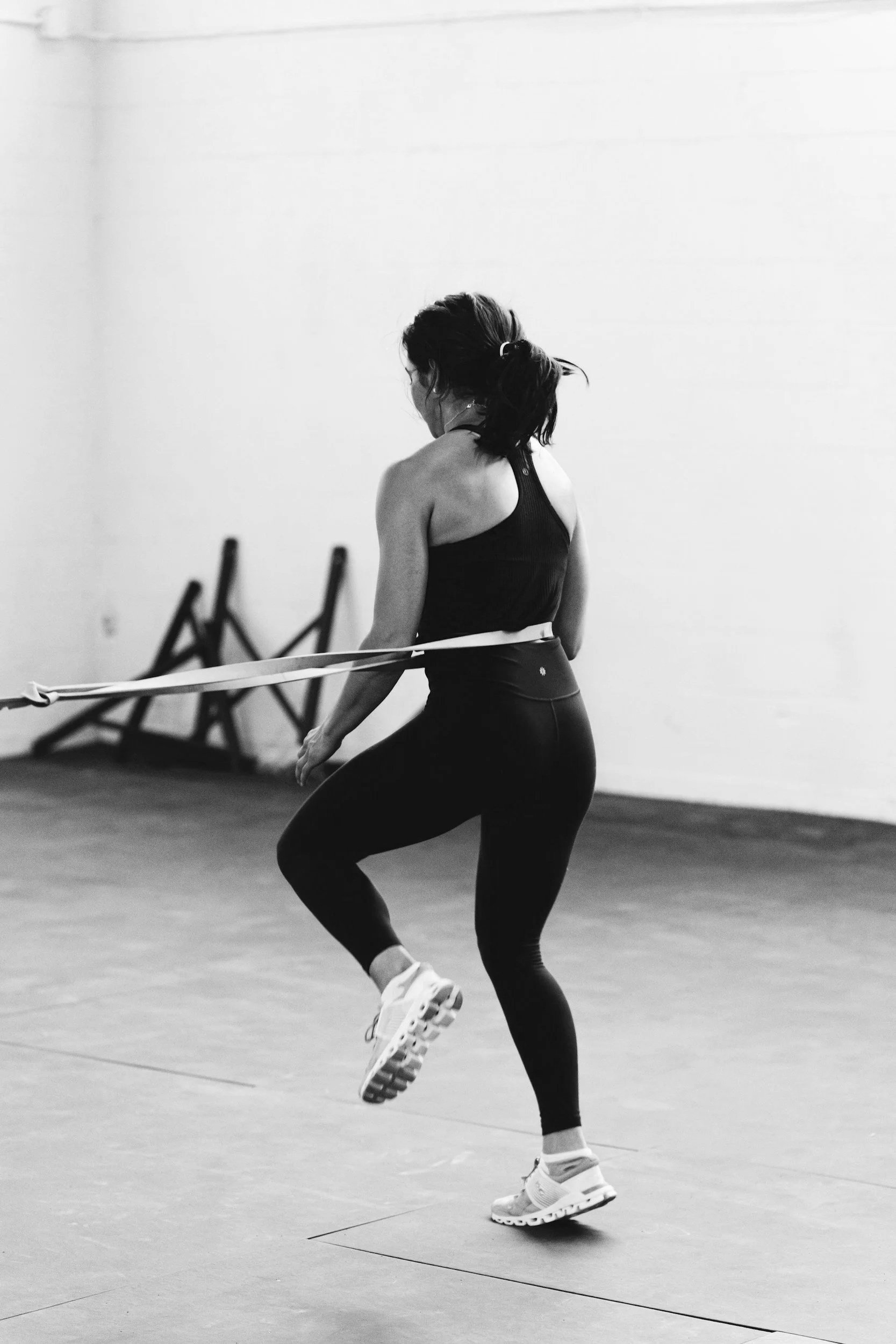 A woman wearing a sports bra and leggings performing a jump with a resistance band in a gym.