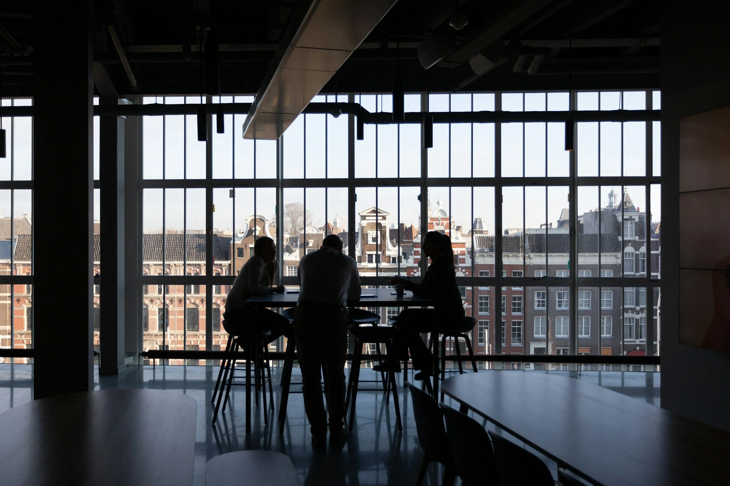Silhouettes of three people sitting at a high table in front of large windows overlooking historic buildings in a city.